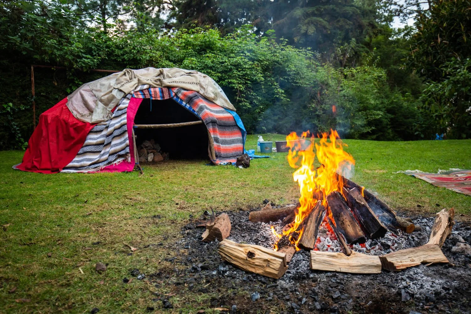 Campfire with bright flames burning logs on a grassy field, with a pile of colorful blankets and a shelter made of blankets and sticks in the background, surrounded by green trees.