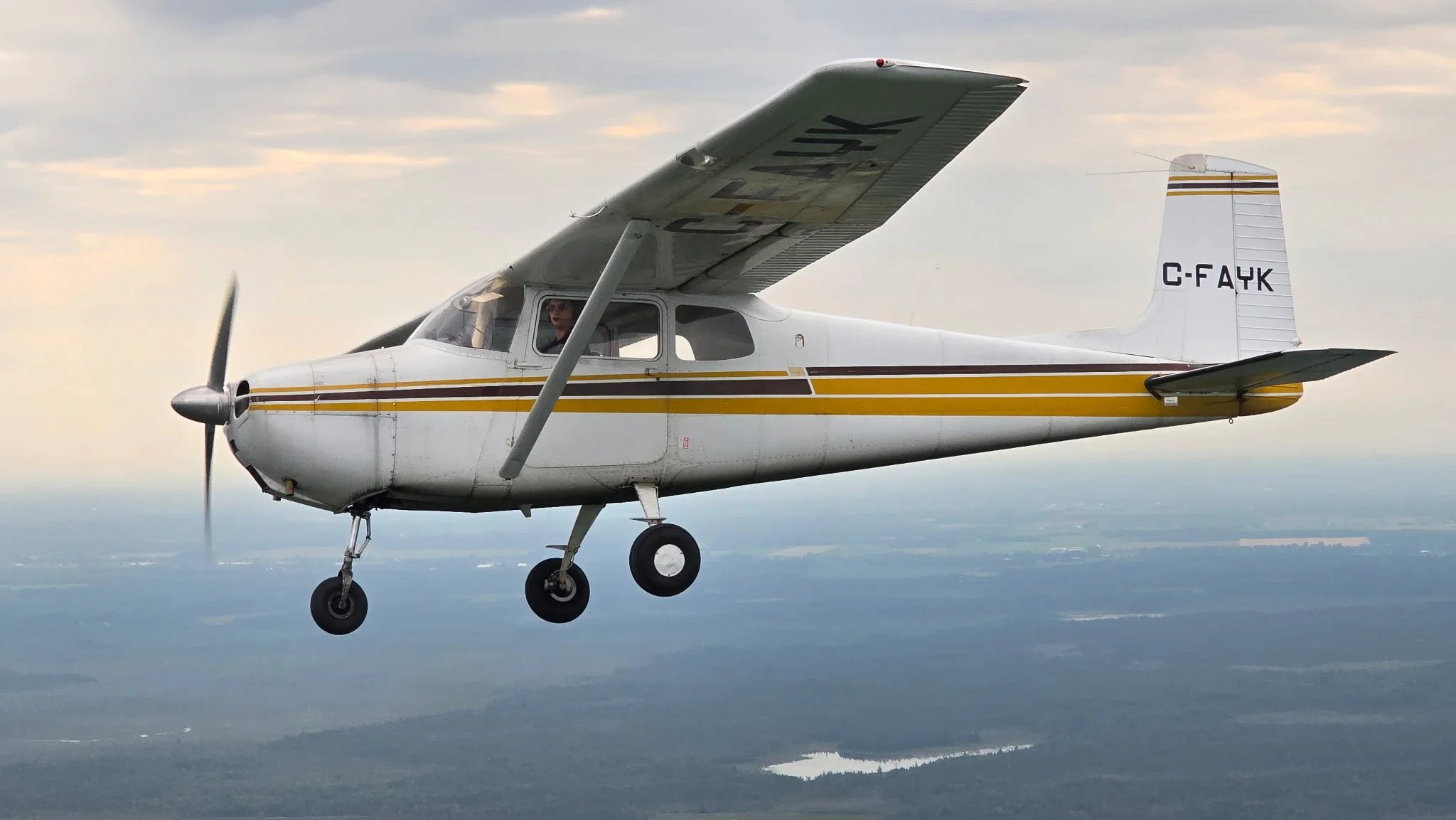 Small single-engine propeller airplane flying through the sky with a cloudy background and a landscape below.
