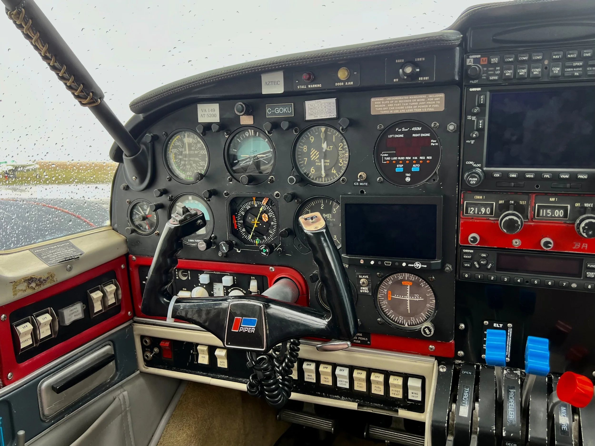 View of an airplane cockpit dashboard with various gauges, switches, and controls, including a PIPER control yoke, during rainy weather outside through the windshield.