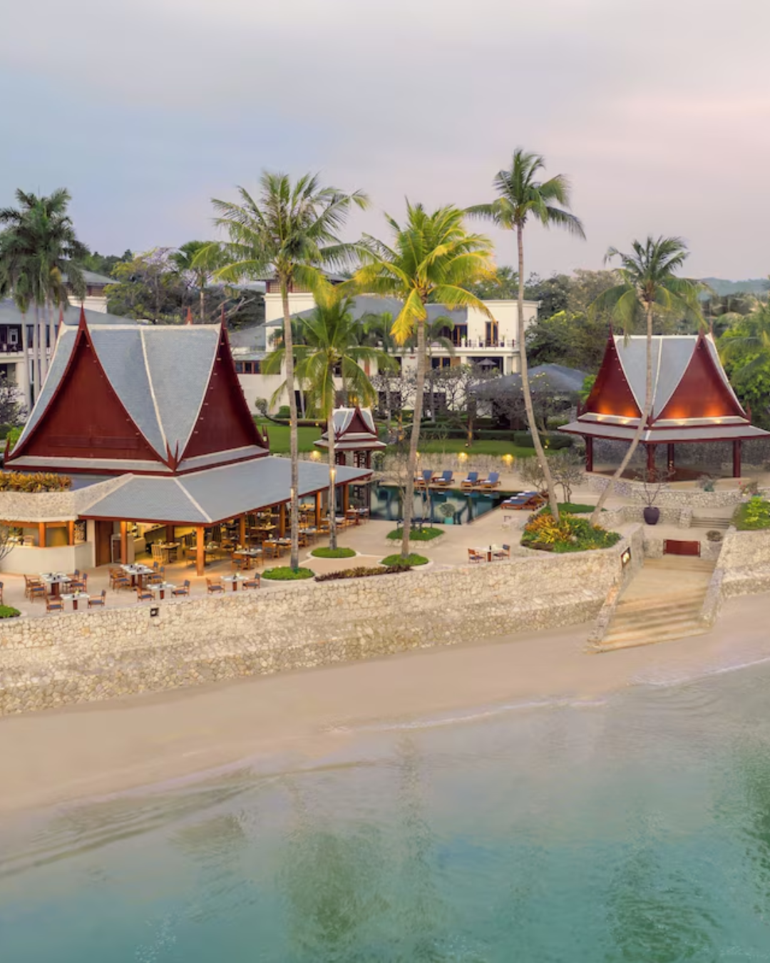 A tropical resort with traditional Thai-style buildings, tall palm trees, a swimming pool, outdoor seating, and beachfront access, during sunset or early evening.