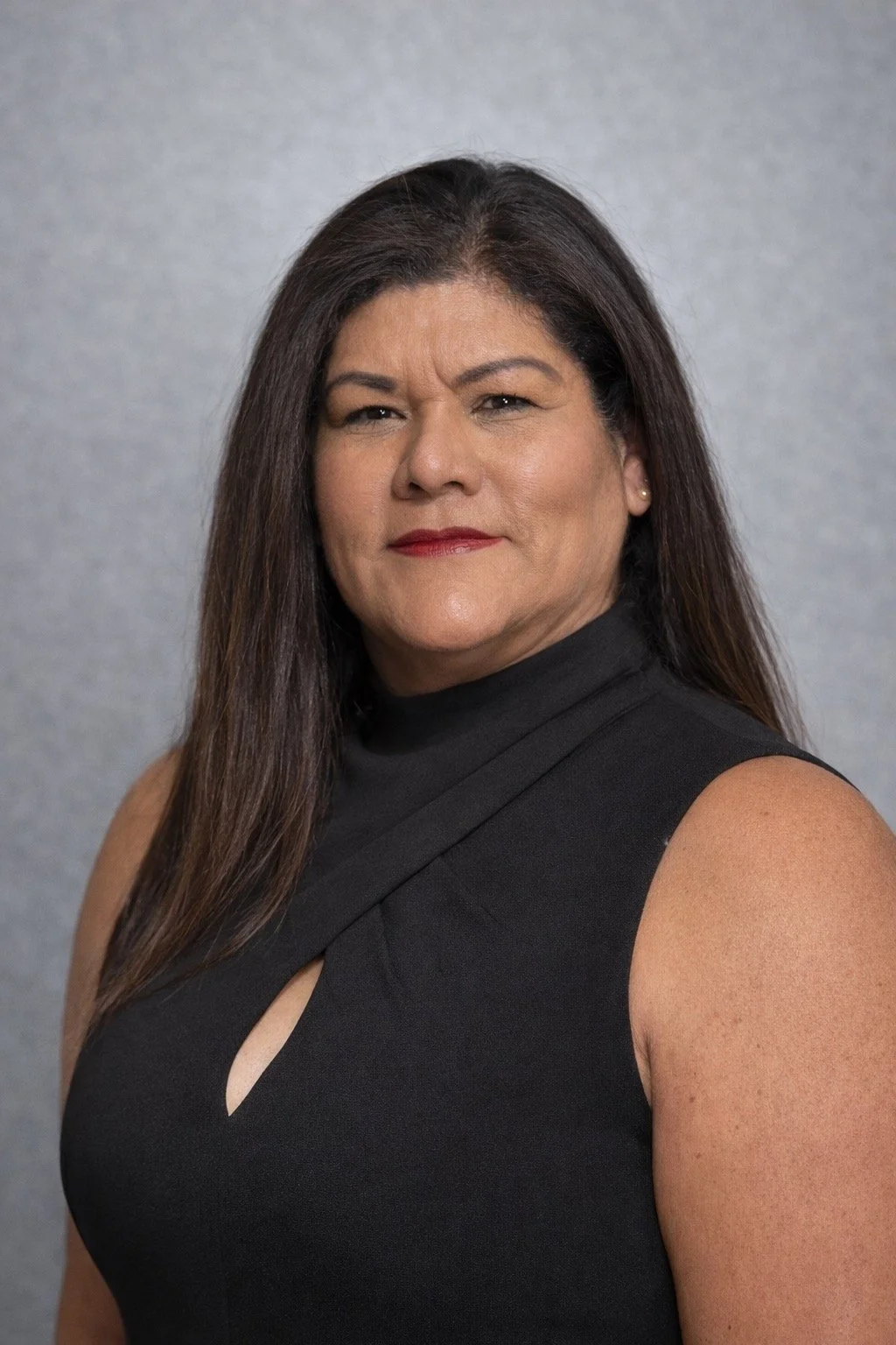 A woman with long dark hair wearing a sleeveless black top with a keyhole detail, looking confidently at the camera against a neutral background.