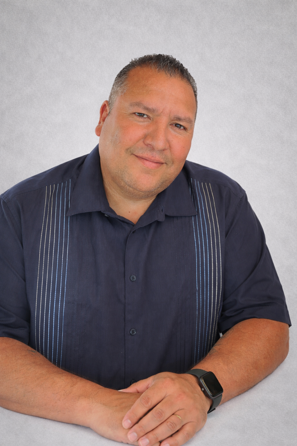 A man with short gray hair, wearing a dark blue shirt with vertical striped patterns, smiling and sitting with his hands folded, against a plain light background.