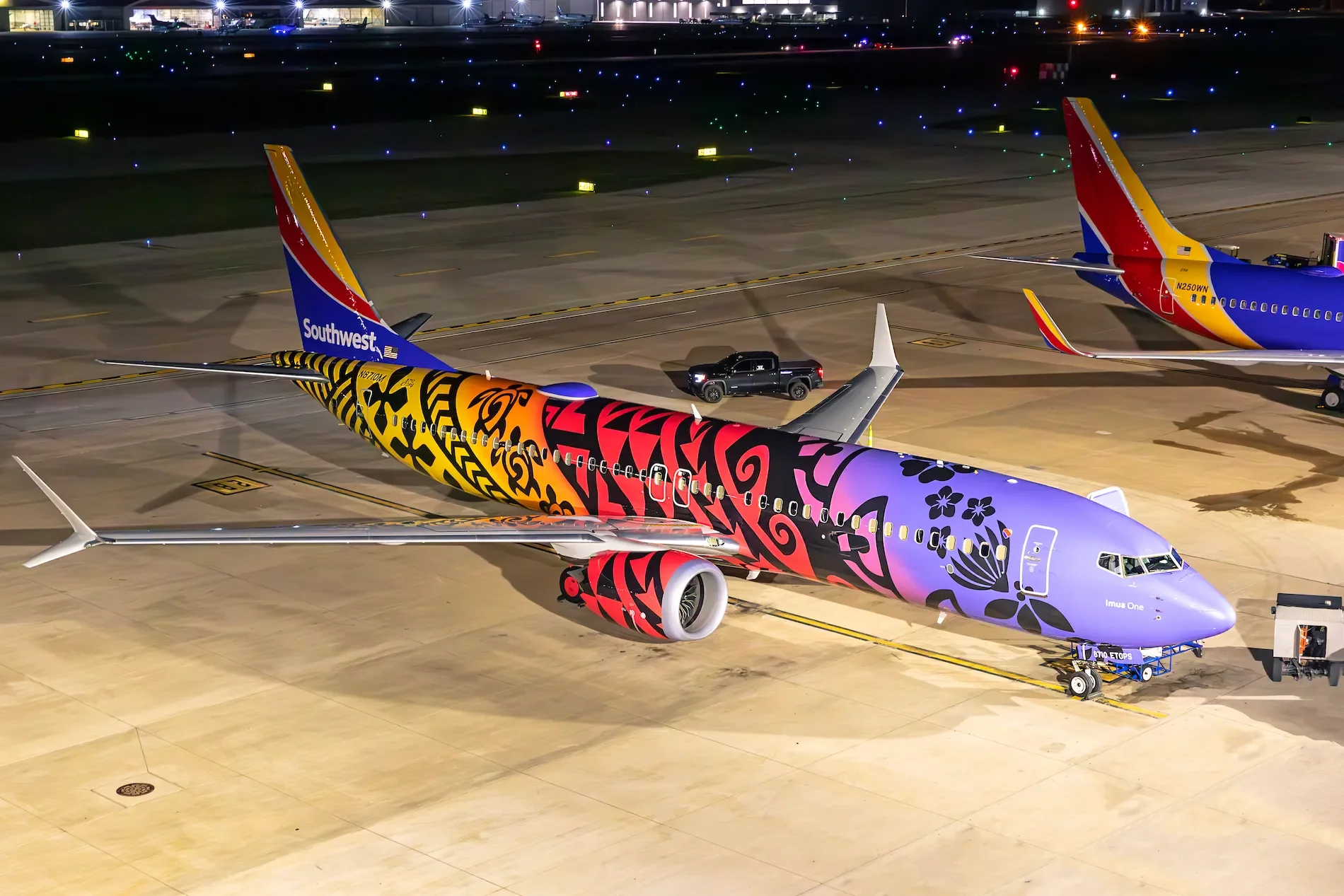 Colorful airplane with floral and tribal designs on the tarmac at night, with two other airplanes painted in Southwest Airlines' red, yellow, and blue airline colors parked nearby, and airport buildings in the background.