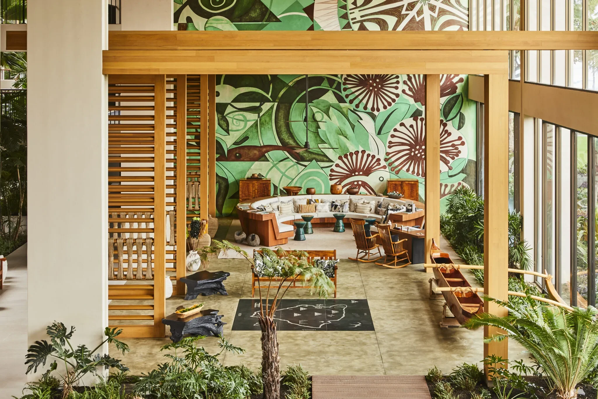 Interior view of a building with a large, colorful mural of abstract plant and animal motifs on the wall. There is a circular seating area with a beige sectional sofa, wooden chairs, and small tables. Large windows let in natural light, and there are numerous green plants and trees inside, creating a lush, natural atmosphere.