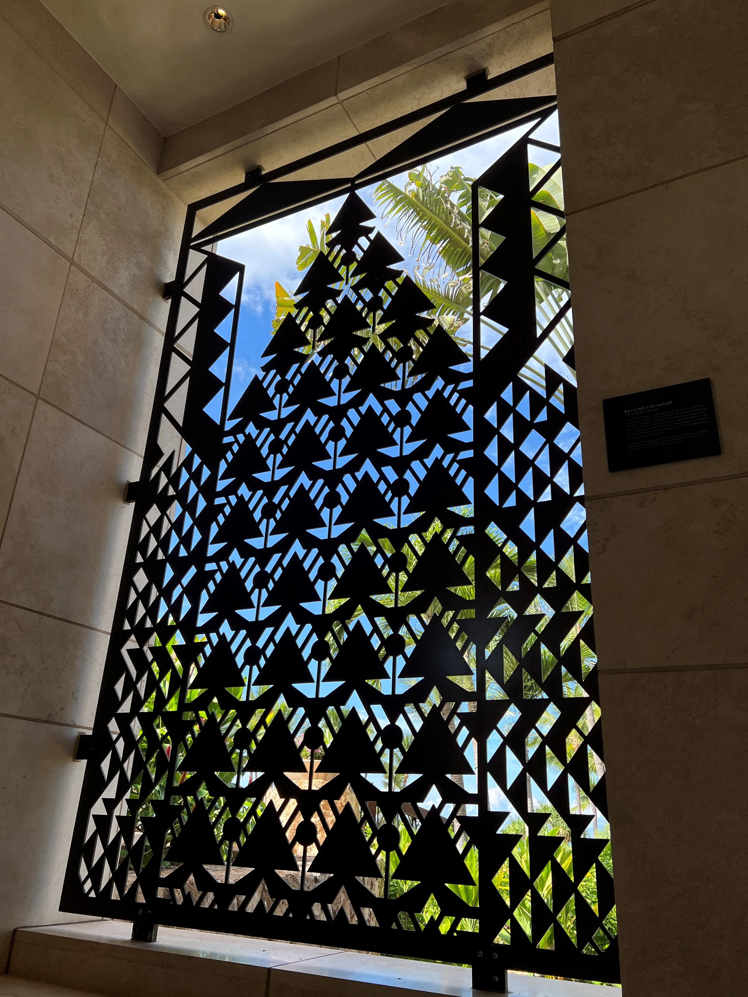 View through a decorative black metal balcony railing, showing a bright blue sky, a few white clouds, and lush green tropical plants.