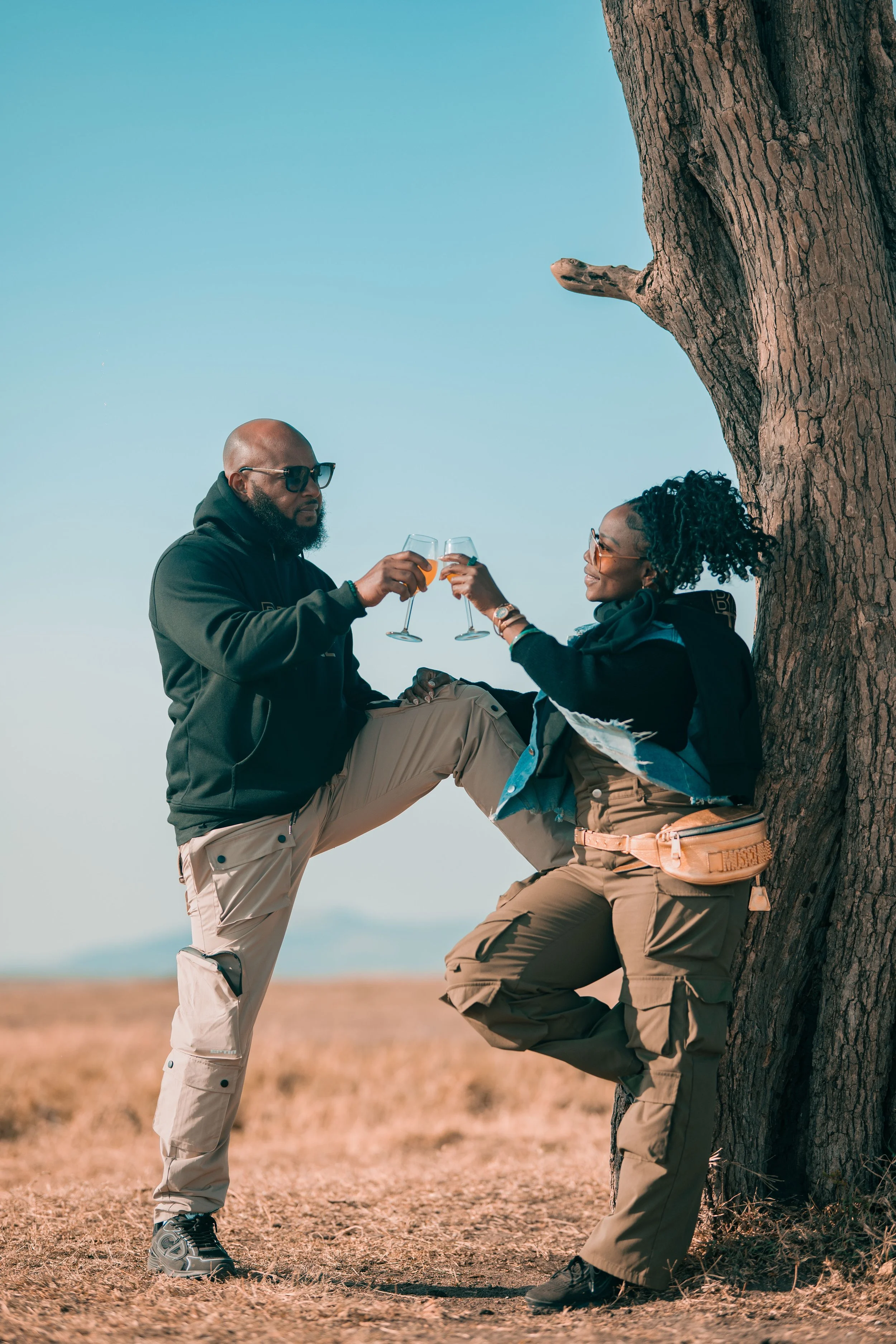 A couple shares a toast beneath a tree in the Maasai Mara, Kenya during an EARTHCANDY TRAVELS journey curated by Jamila Crawford Pécou, surrounded by expansive landscapes that reflect romance, connection, and unforgettable cultural travel experiences