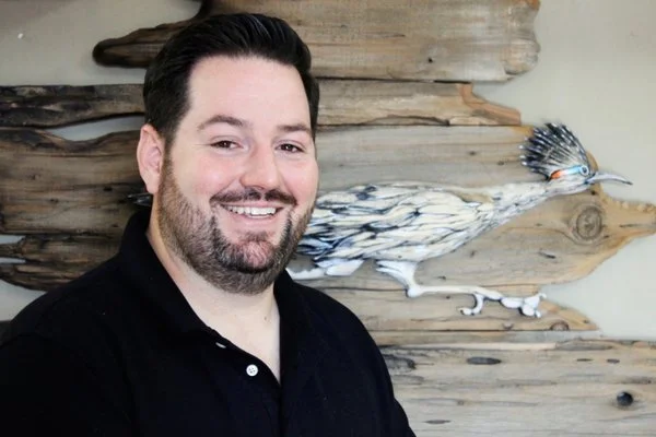 A smiling man with dark hair and a beard wearing a black polo shirt stands in front of a rustic wooden wall with a decorative bird sculpture.
