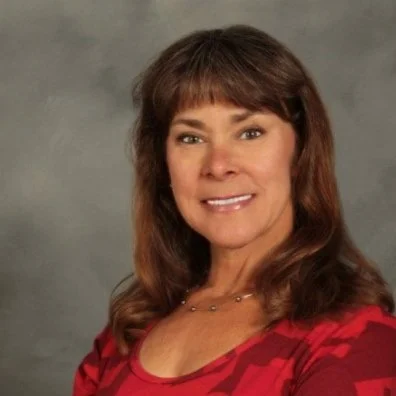 A woman with brown hair wearing a red patterned top smiling at the camera against a gray background.
