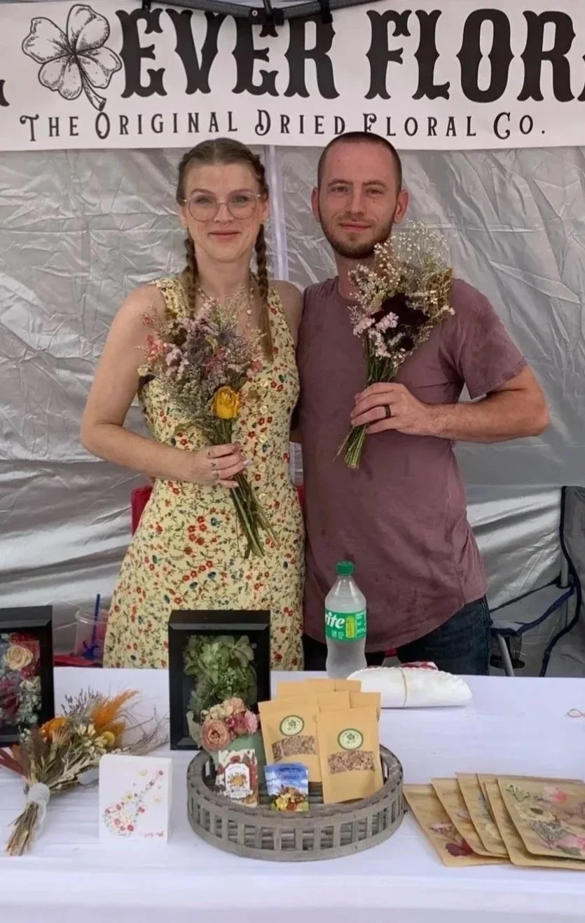 A man and woman stand side by side, each holding a bouquet of flowers, smiling at the camera. They are under a tent with a sign that says 'The Original Dried Floral Co.' on the banner behind them. The table in front of them has decorative items, framed floral arrangements, and packets of dried flowers.