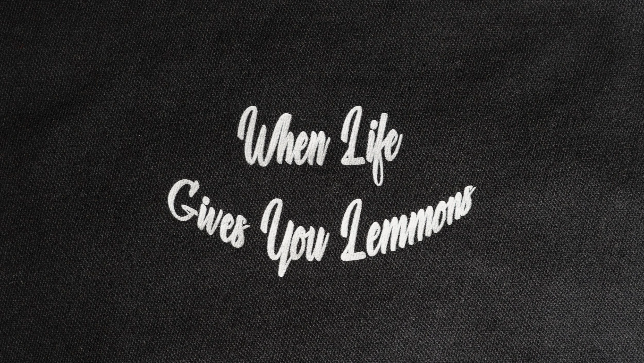 Closeup shot of plain black t-shirt. Features white text that reads ‘When Life Gives You Lemmon’