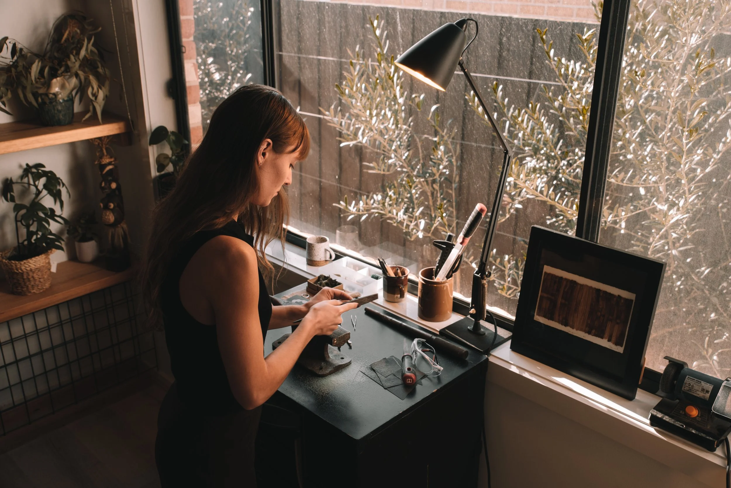 A woman standing at a black work desk by a window, looking at her phone, surrounded by art supplies, a desk lamp turned on, and a small TV or monitor on the desk.