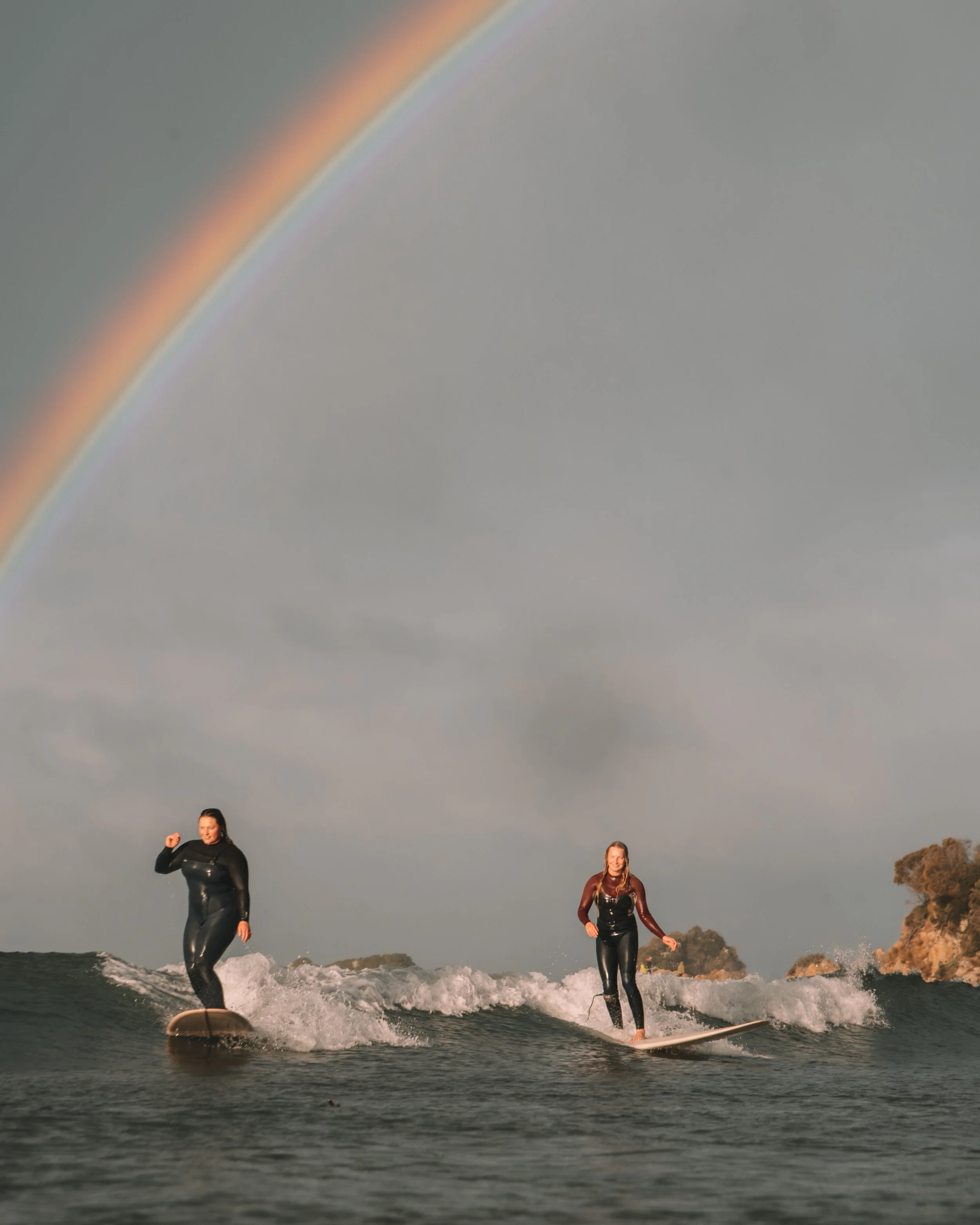 Two women surfing in the ocean under a rainbow with cloudy skies and rocky coastline in the background.