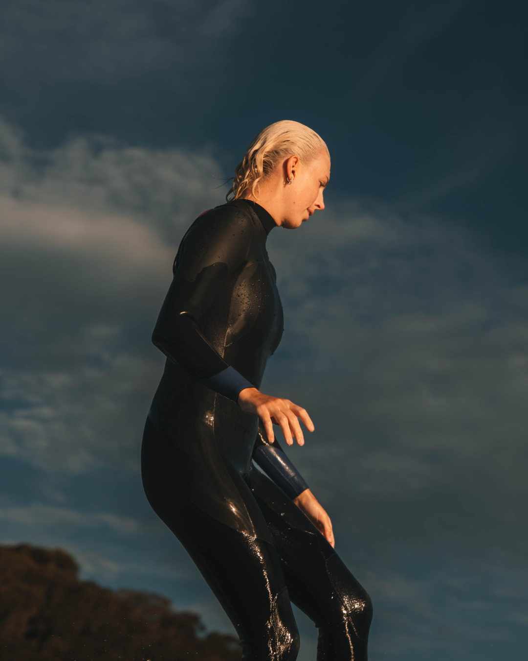 A person in a black wet suit standing outdoors against a cloudy sky, with water droplets on the suit.