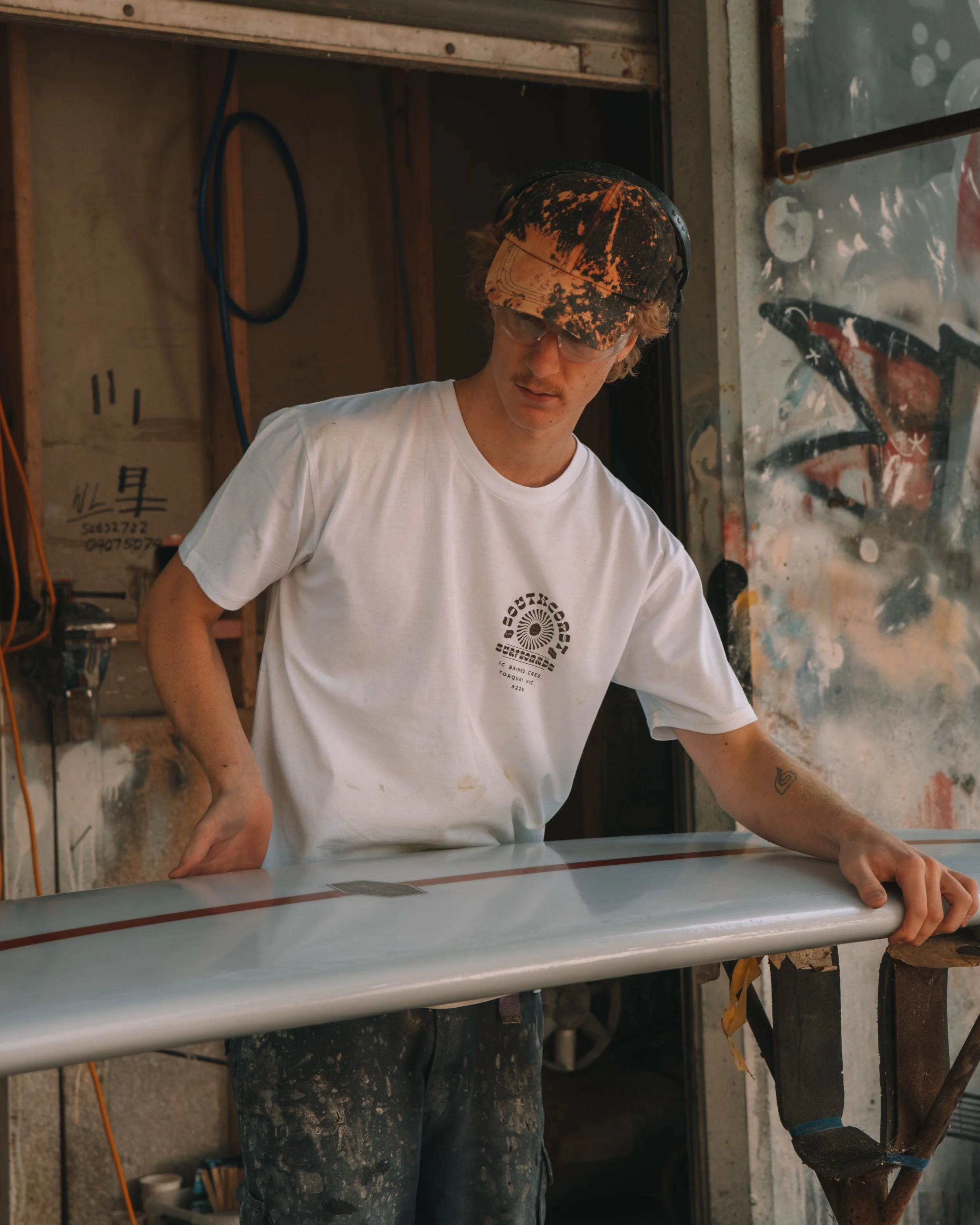 A young man wearing a white t-shirt, a leather cap, and safety glasses, standing beside a surfboard in a workshop.