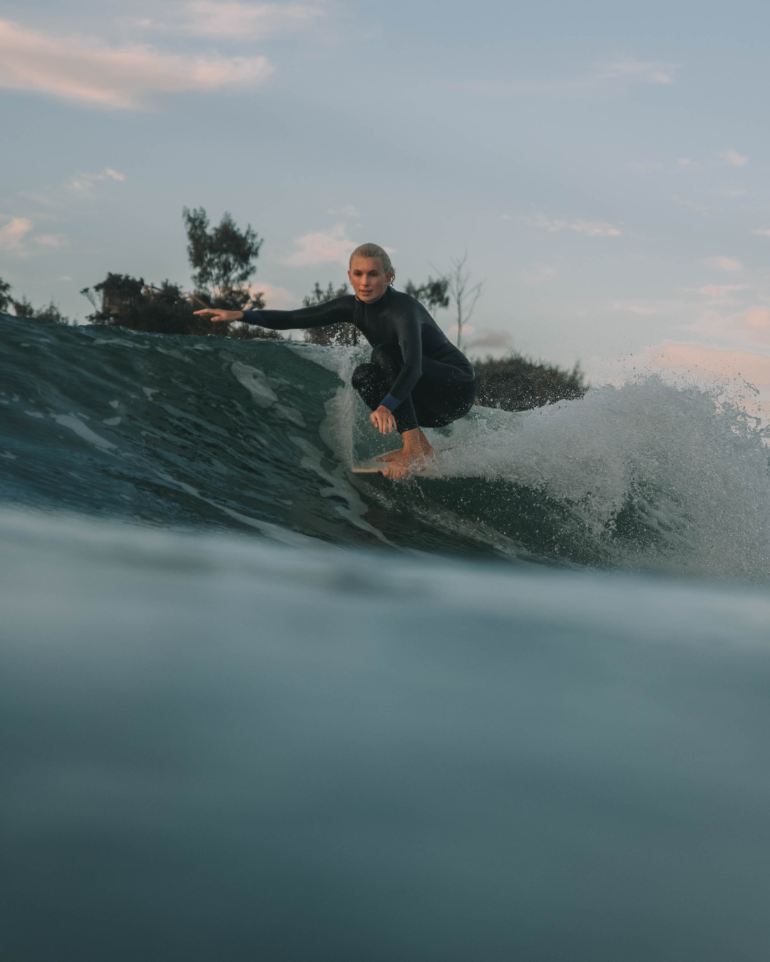 A woman in a black wetsuit surfing on a wave in the ocean during late afternoon or early evening.
