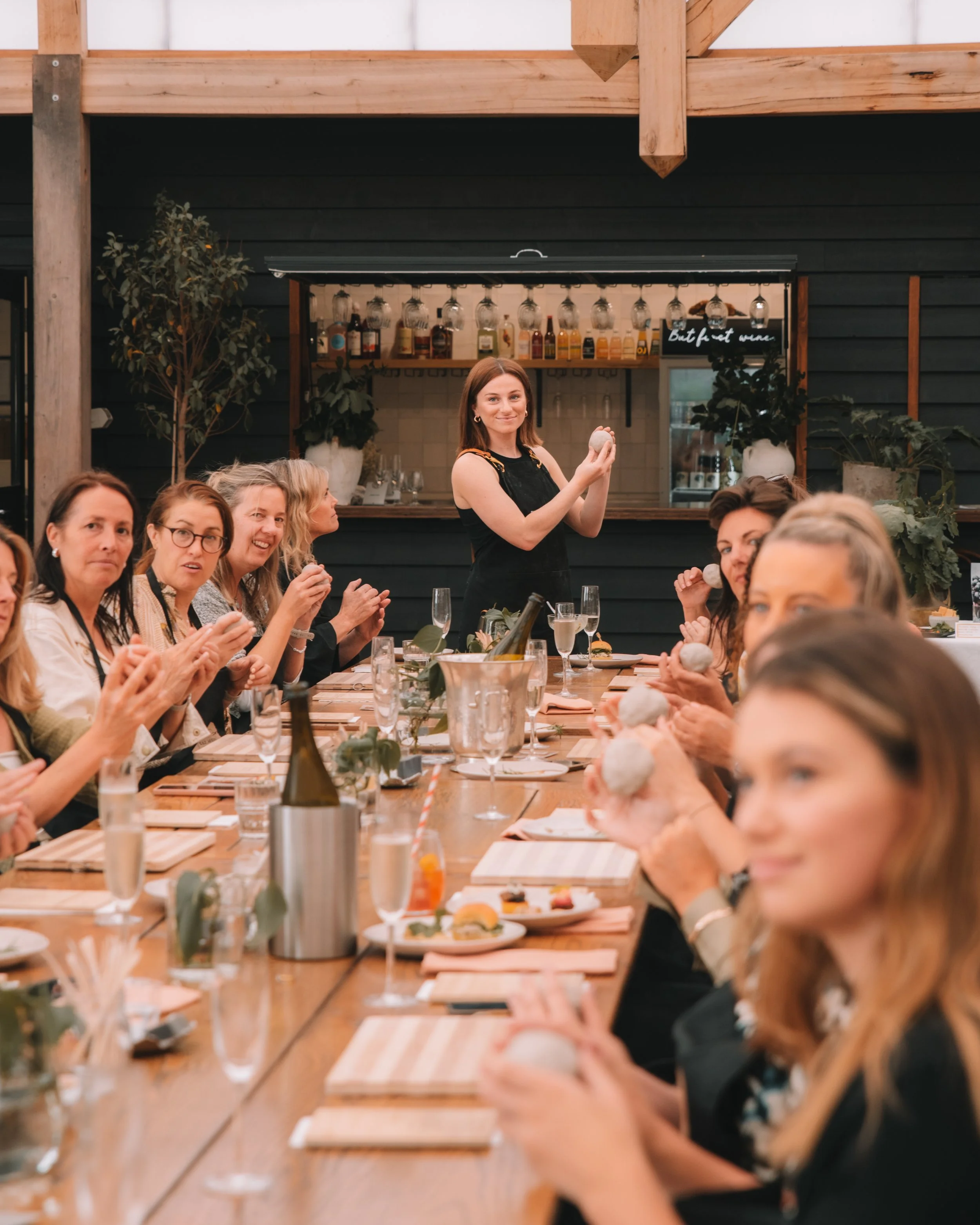 A group of women seated at a long dining table, clapping and smiling, with a woman standing at the end holding a ball, in a restaurant setting with a bar in the background.