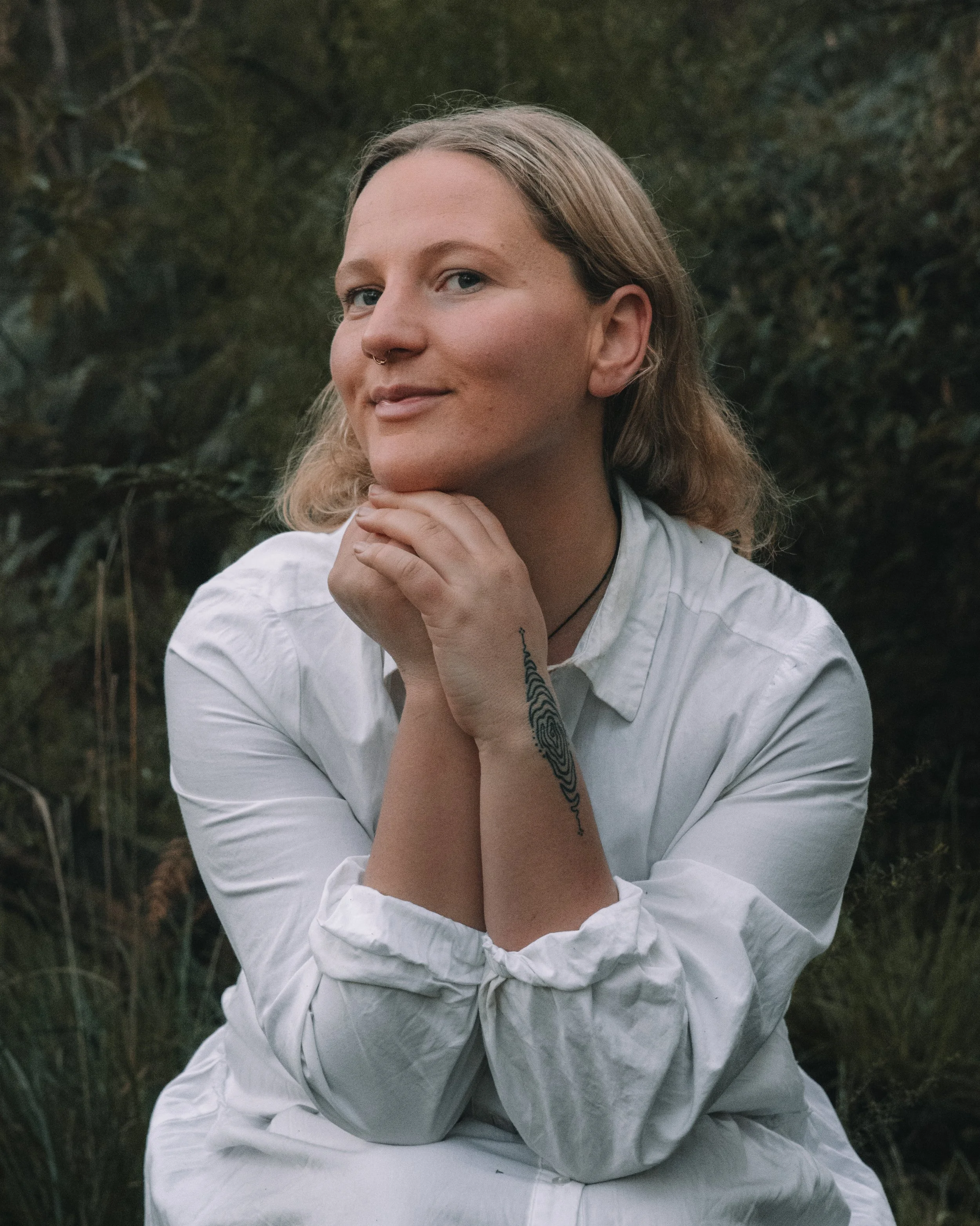 A woman with blonde hair, wearing a white shirt, sitting outdoors with greenery in the background, resting her chin on her hands, smiling slightly.