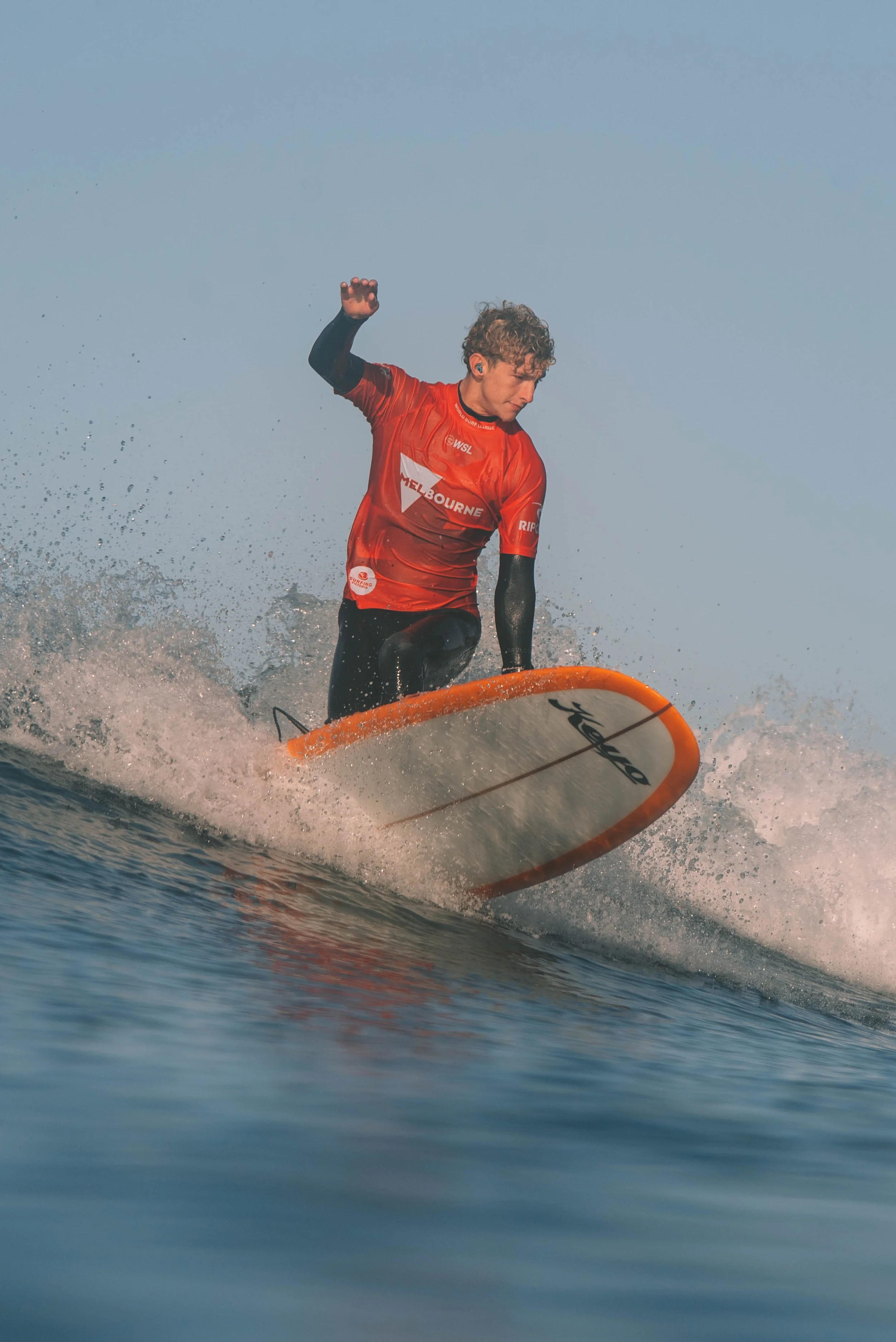 Surfer riding a wave on a sunny day, wearing a red wetsuit top with 'Melbourne' written on it, black wetsuit pants, and balancing on an orange surfboard.