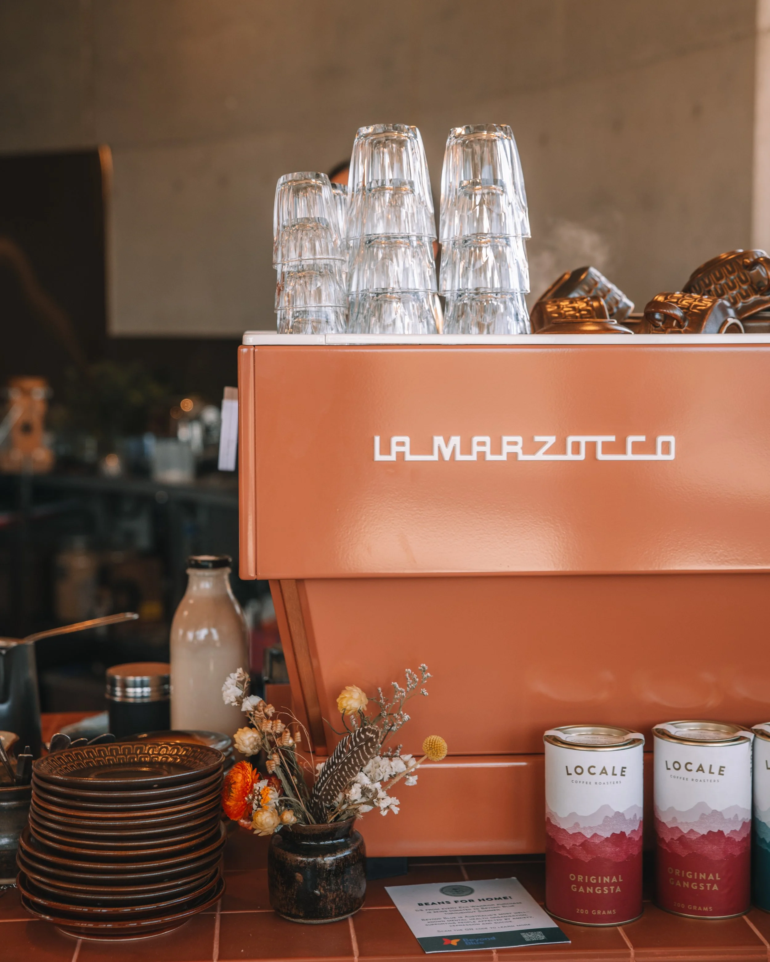 A vintage orange espresso machine with glasses stacked upside down on top, canned coffee labeled 'Locale' stacked on the side, plates, a bottle, a small bouquet of flowers in a vase, and a small permission note on a countertop.