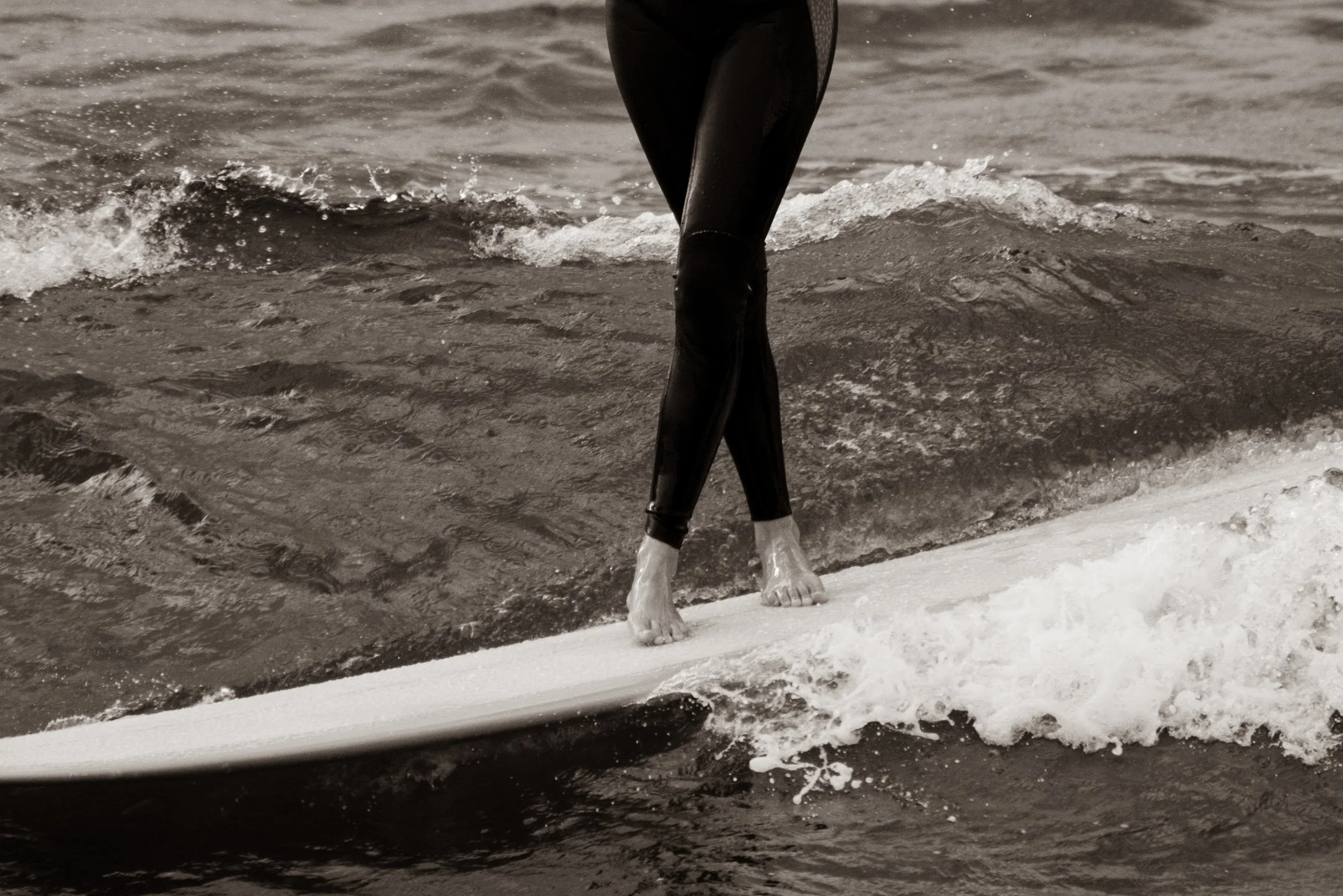 A person in a wetsuit standing on a paddleboard on the water, with their legs crossed at the ankles, legs and hands visible, part of the paddleboard visible in the water.