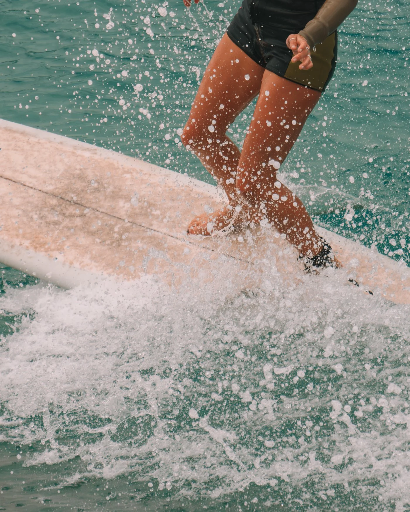 Person in black swim shorts balancing on a surfboard in water, splashing water around.