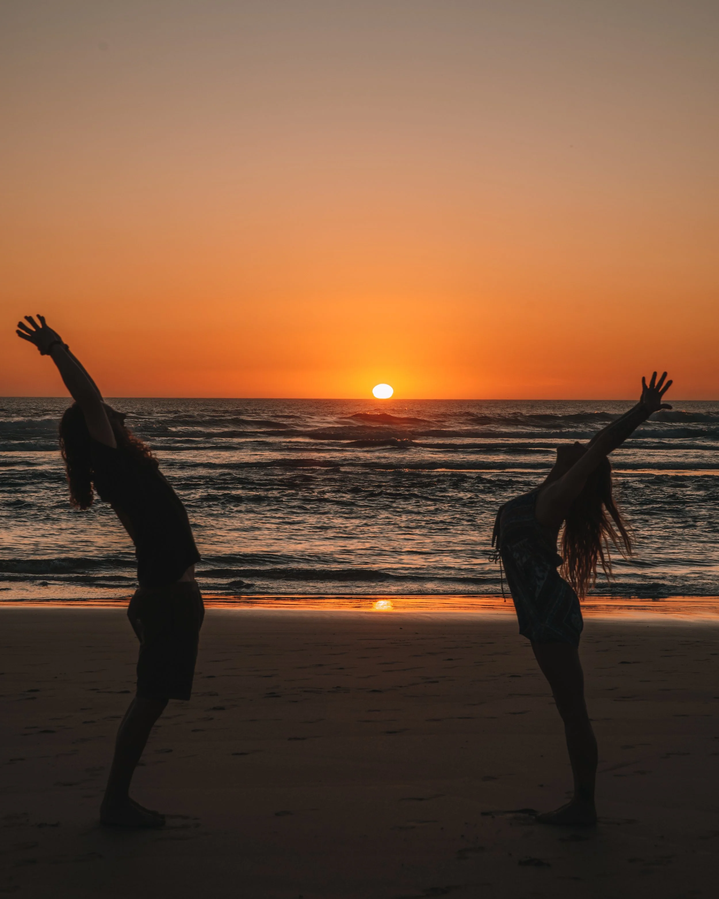 Two women dancing on a beach at sunset with the sun setting over the ocean in the background.