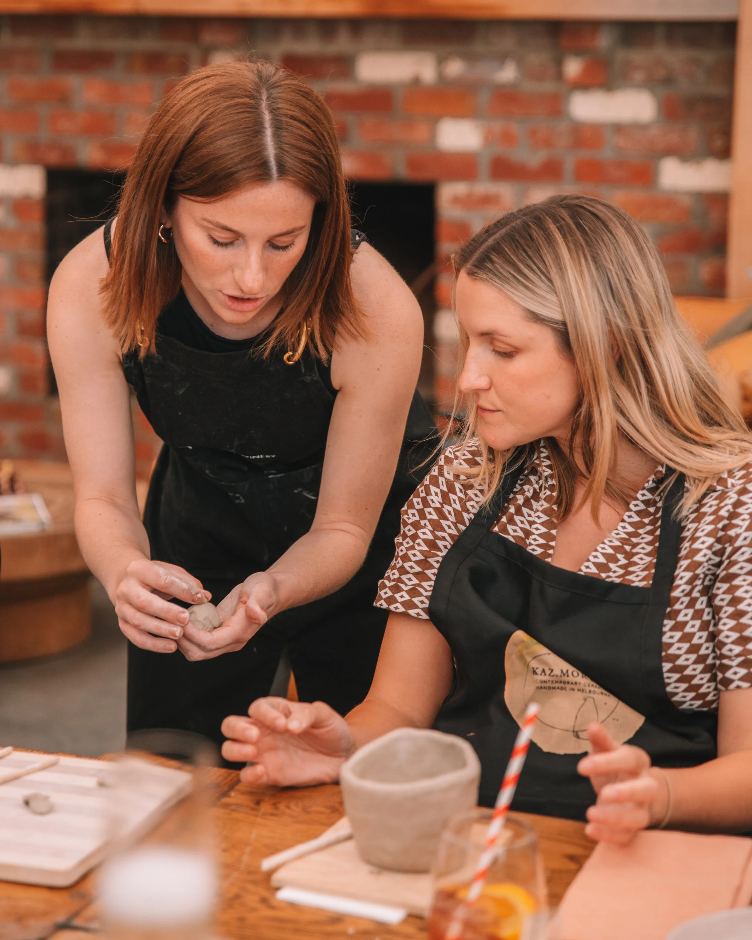 Two women making pottery at a table, one helping the other design a clay piece, in a cozy workshop with exposed brick wall.
