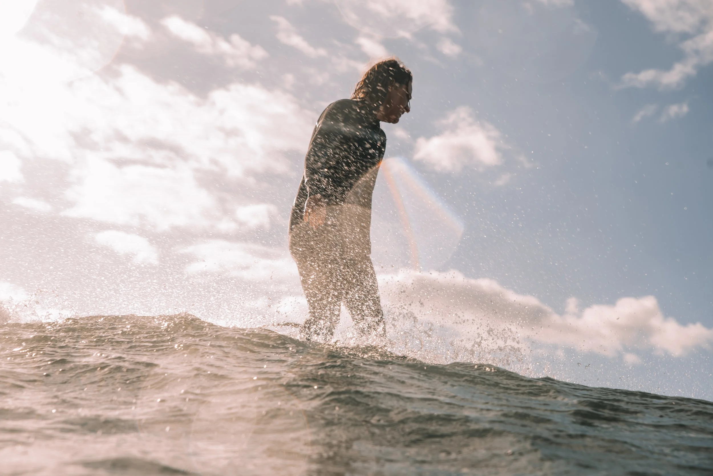 Person standing in the water on a surfboard, splashing water with the sky and clouds in the background.