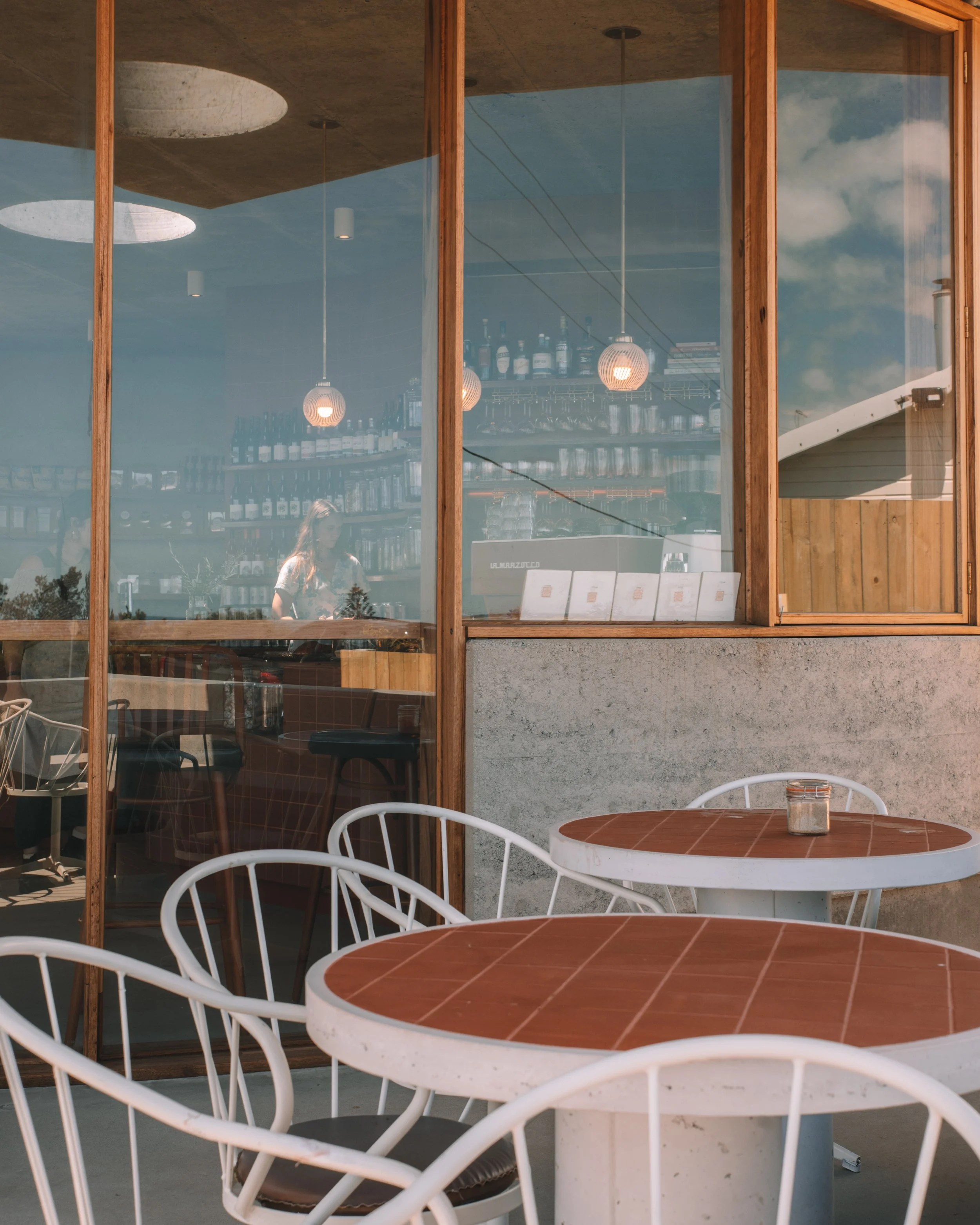 An outdoor patio with brown round tables and white chairs in front of a glass window showing a bar and a person inside.