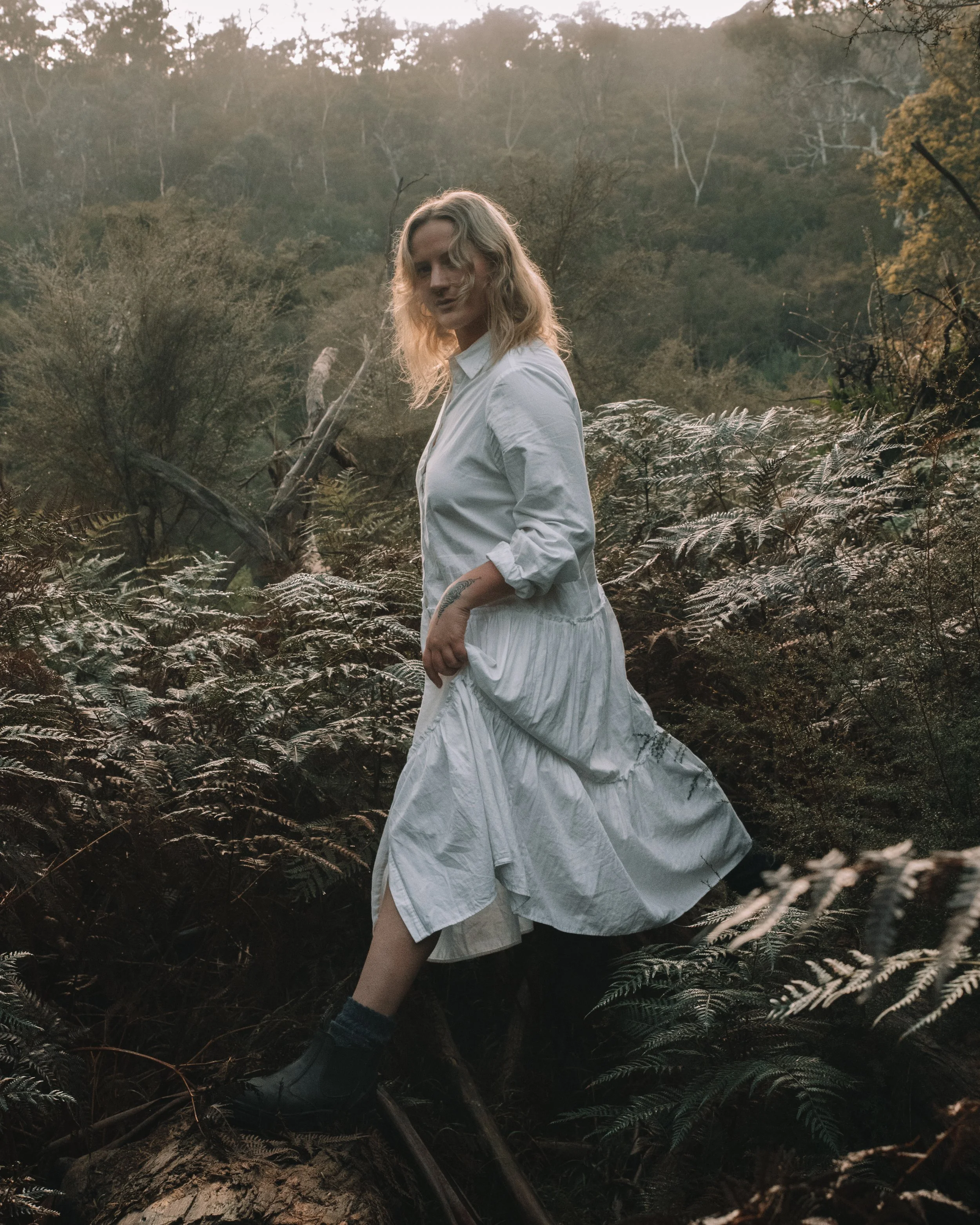 A woman in a white dress and dark boots standing amidst ferns in a forest at sunset.