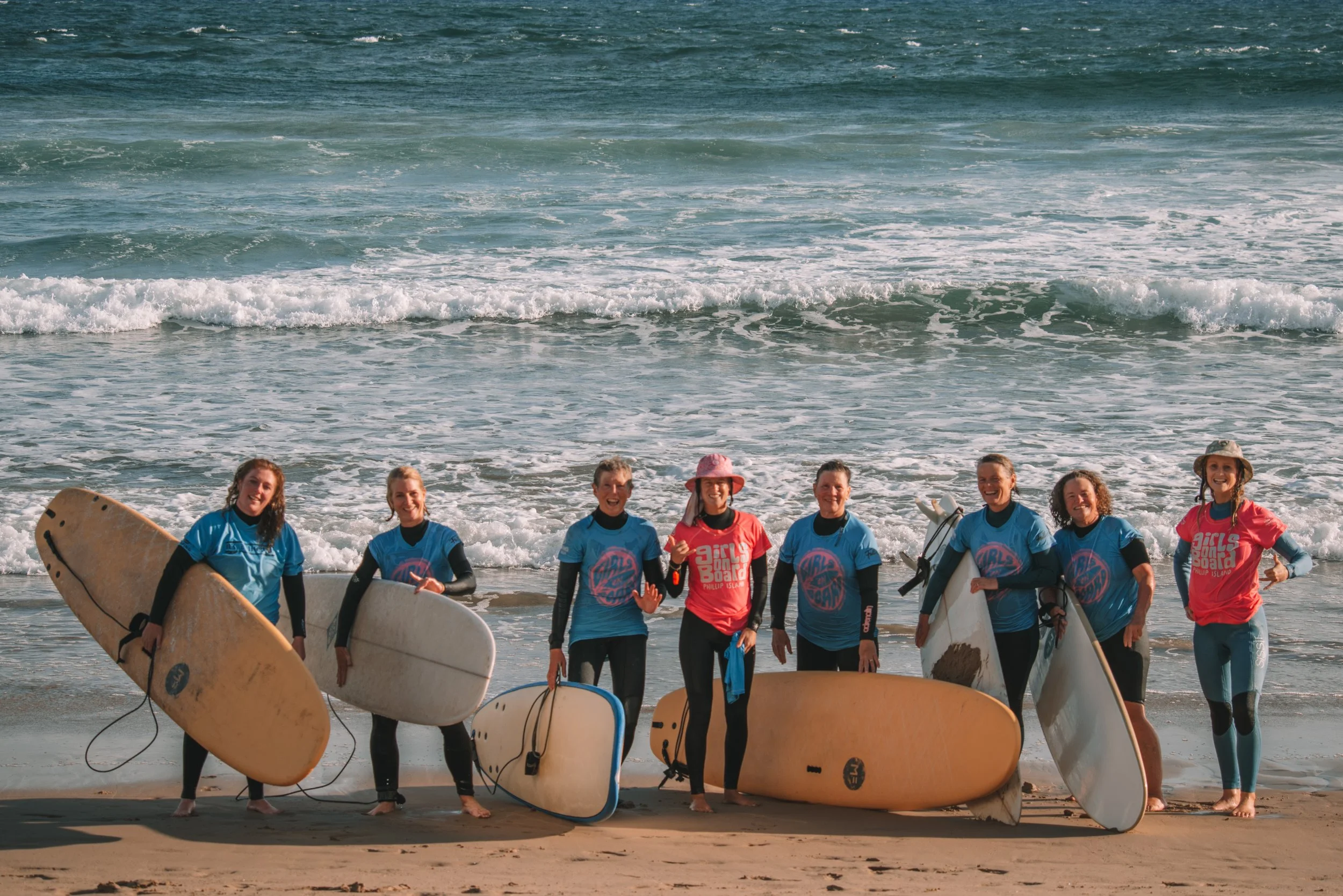 Group of women in wetsuits and rash guards standing on the beach holding surfboards, smiling after surfing session with ocean waves in background.
