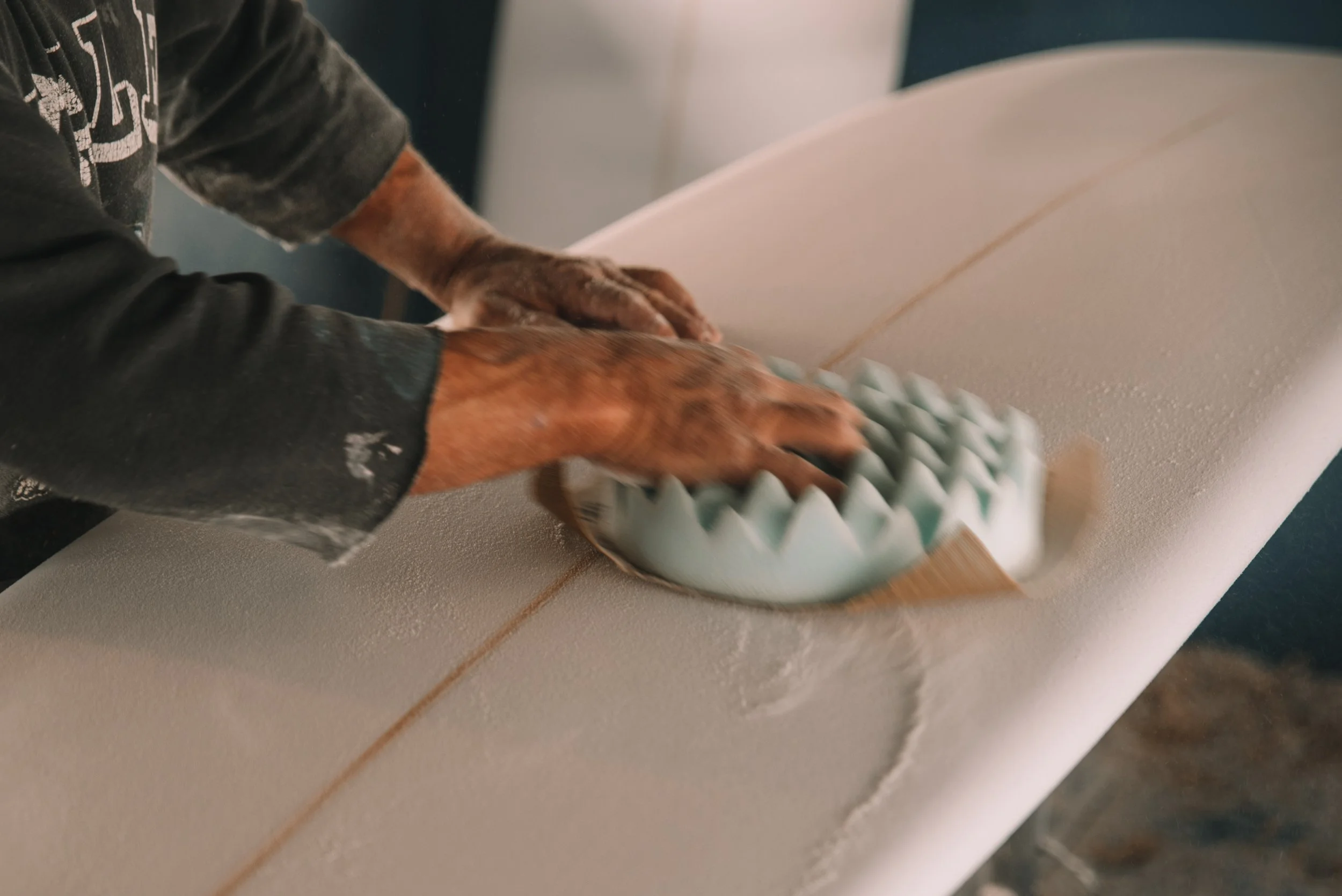 Hands shaping a surfboard with a shaping tool inside a rail. The person is wearing a black long sleeve shirt with white lettering and has some sanding dust on their hands and shirt.
