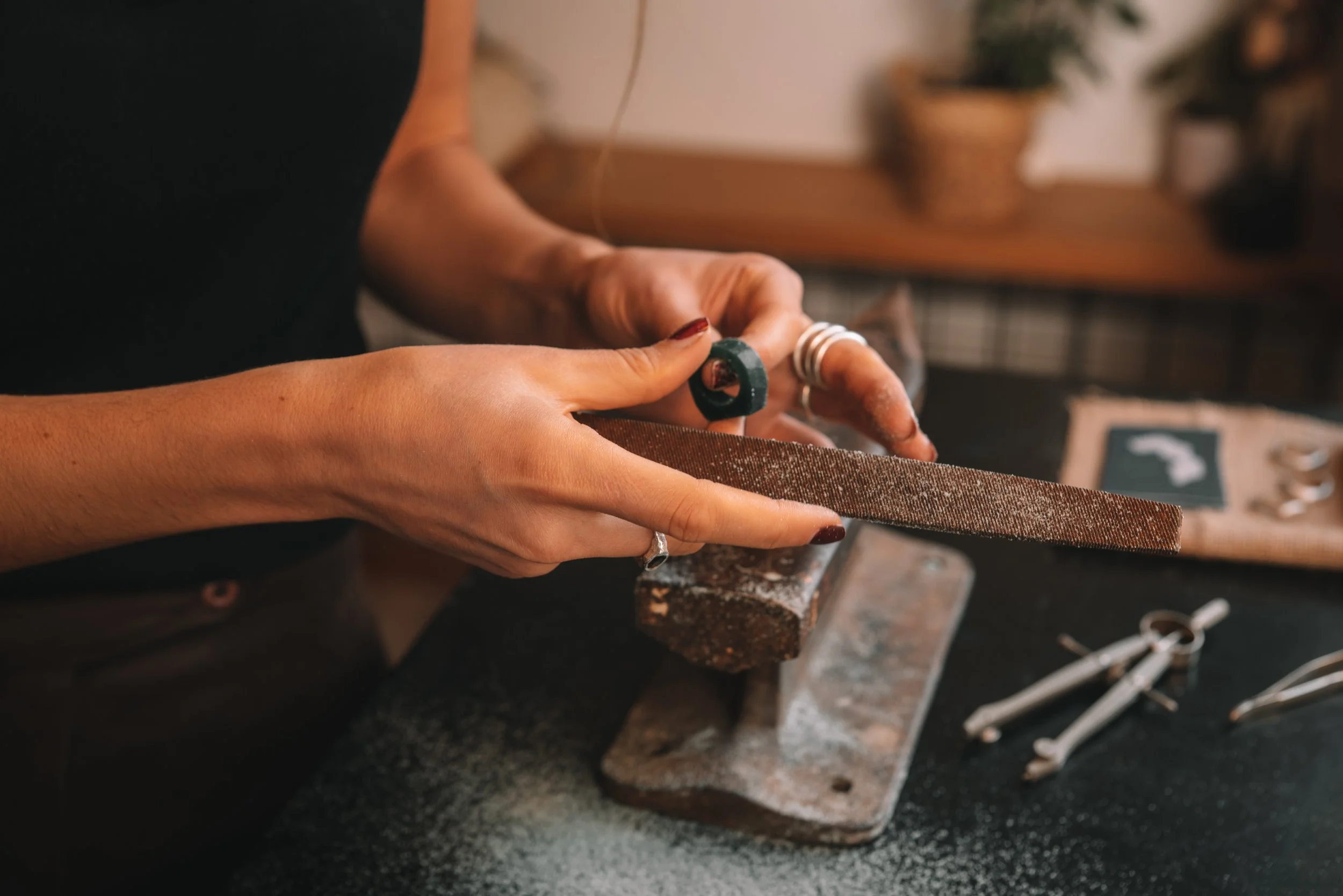 Person polishing jewelry with a file and polishing tool on a workbench surrounded by jewelry-making tools.