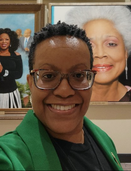 A woman smiles and wears a green blazer. She is standing in front of paintings in an art gallery.