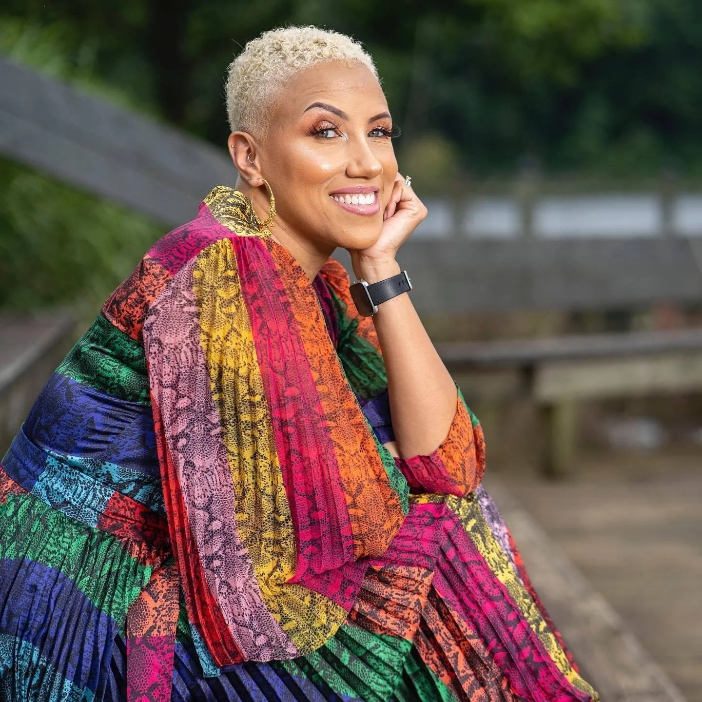 A woman sits at an angle and smiles at the camera. She has short blonde hair and is wearing a brightly colored jumpsuit.