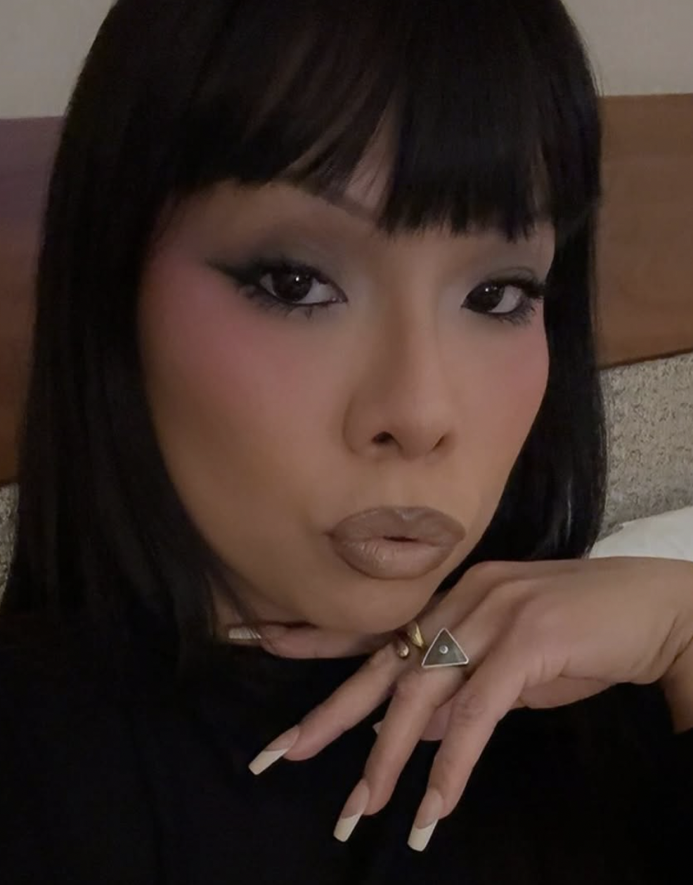 Close-up portrait of a medium-complexioned Native woman with dark straight hair and bangs, wearing makeup with bold eyeliner and nude lipstick, resting her chin on her hand with geometric rings, in a dimly lit room.