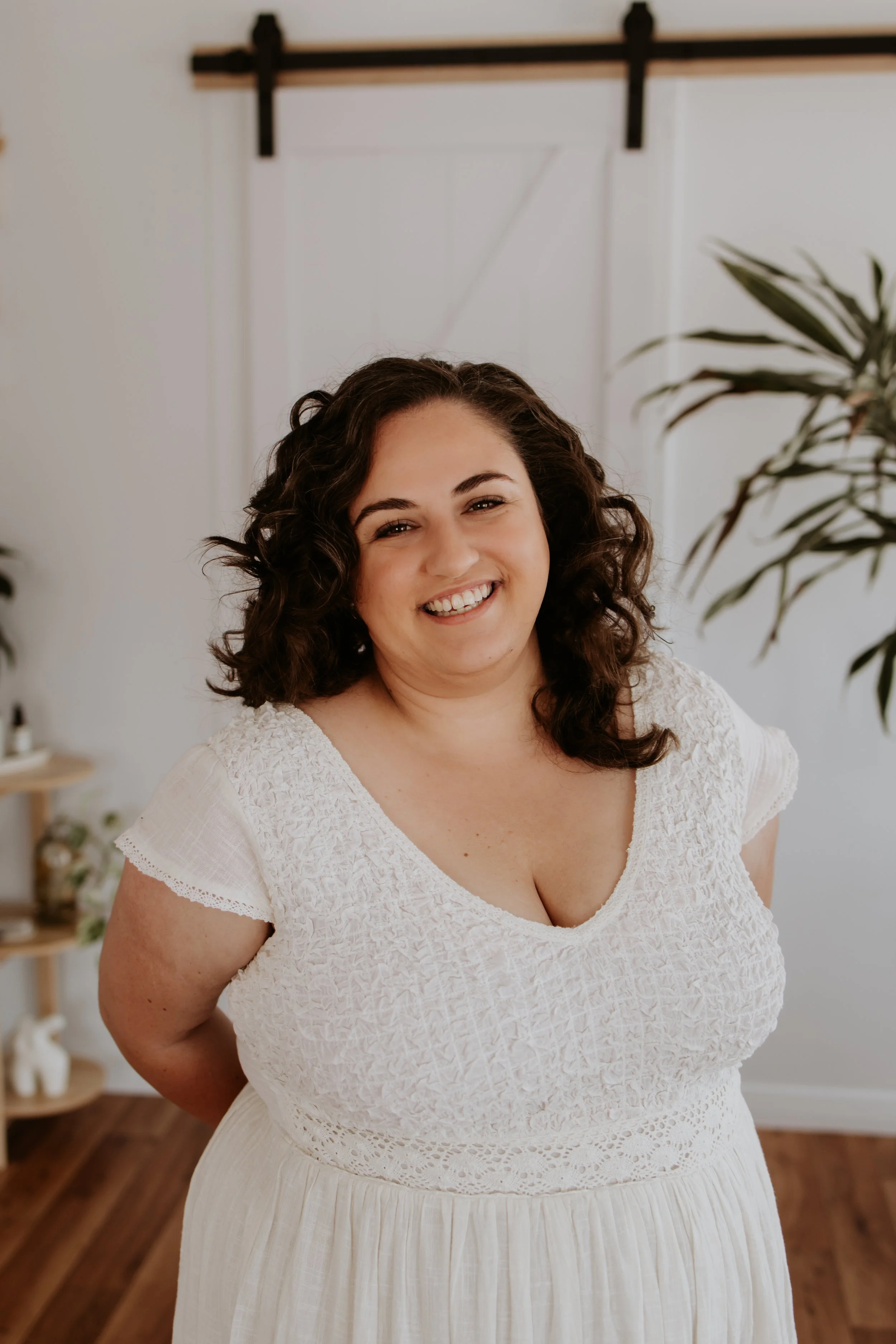 Smiling woman with curly hair wearing a white dress in a bright room with plants and wooden floor.