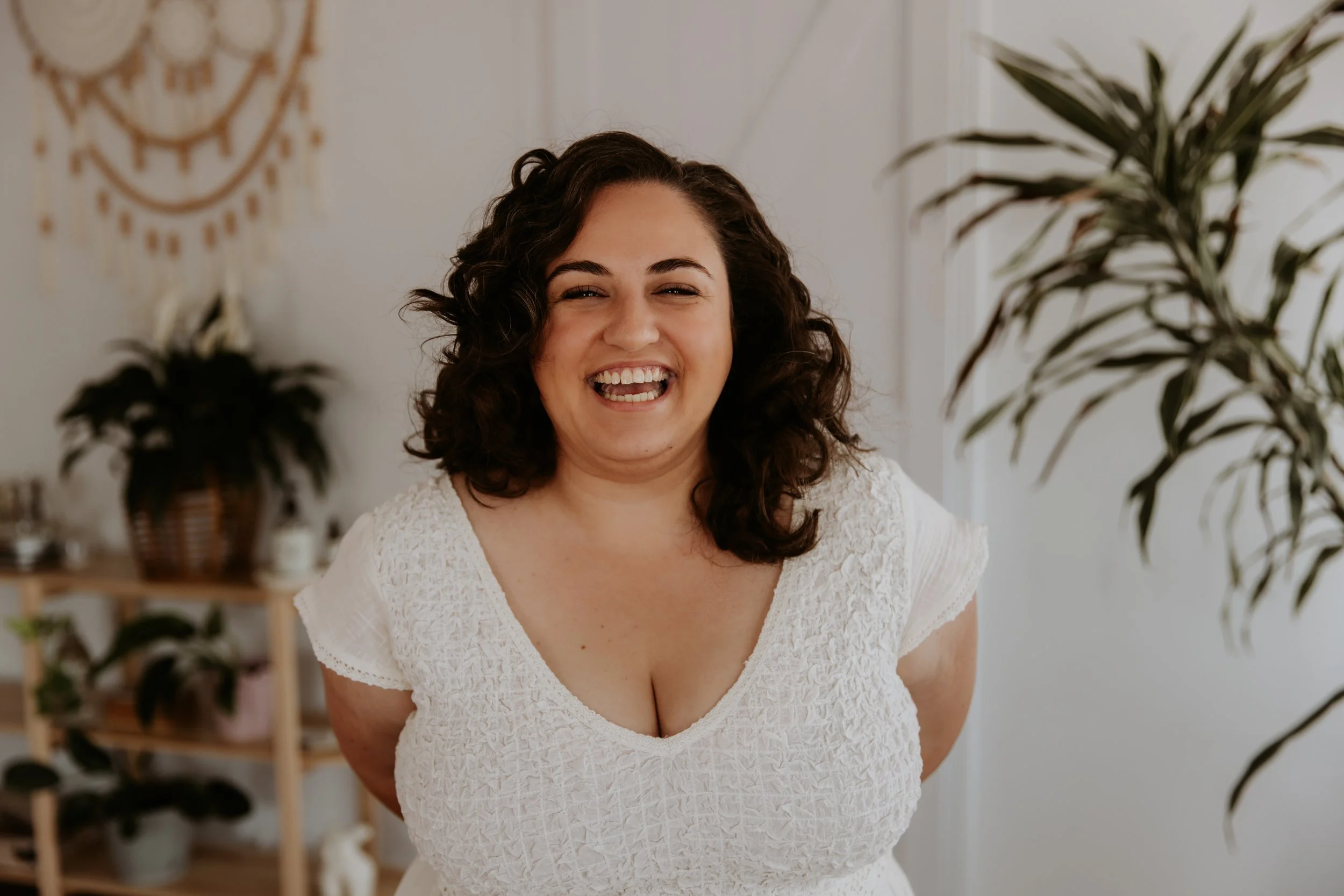 A woman with curly hair is smiling broadly, standing in a room with plants and a decorative wall hanging.