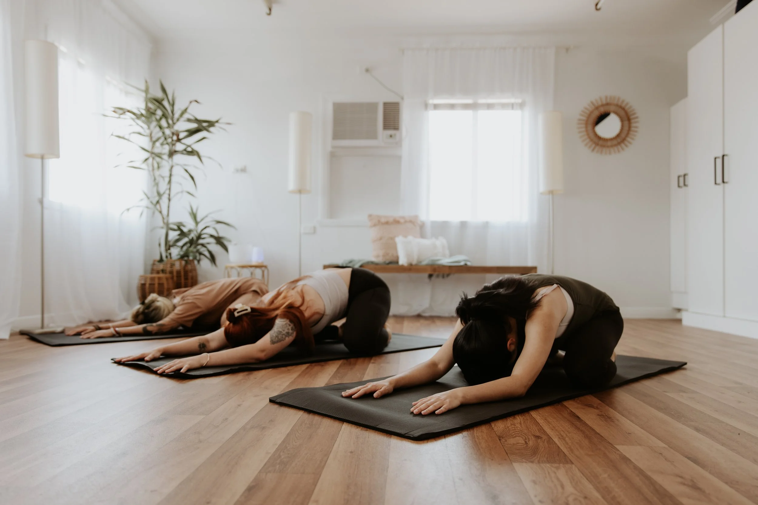 Three women practicing yoga in child's pose on mats in a bright, minimalist room with wooden floors and indoor plants.
