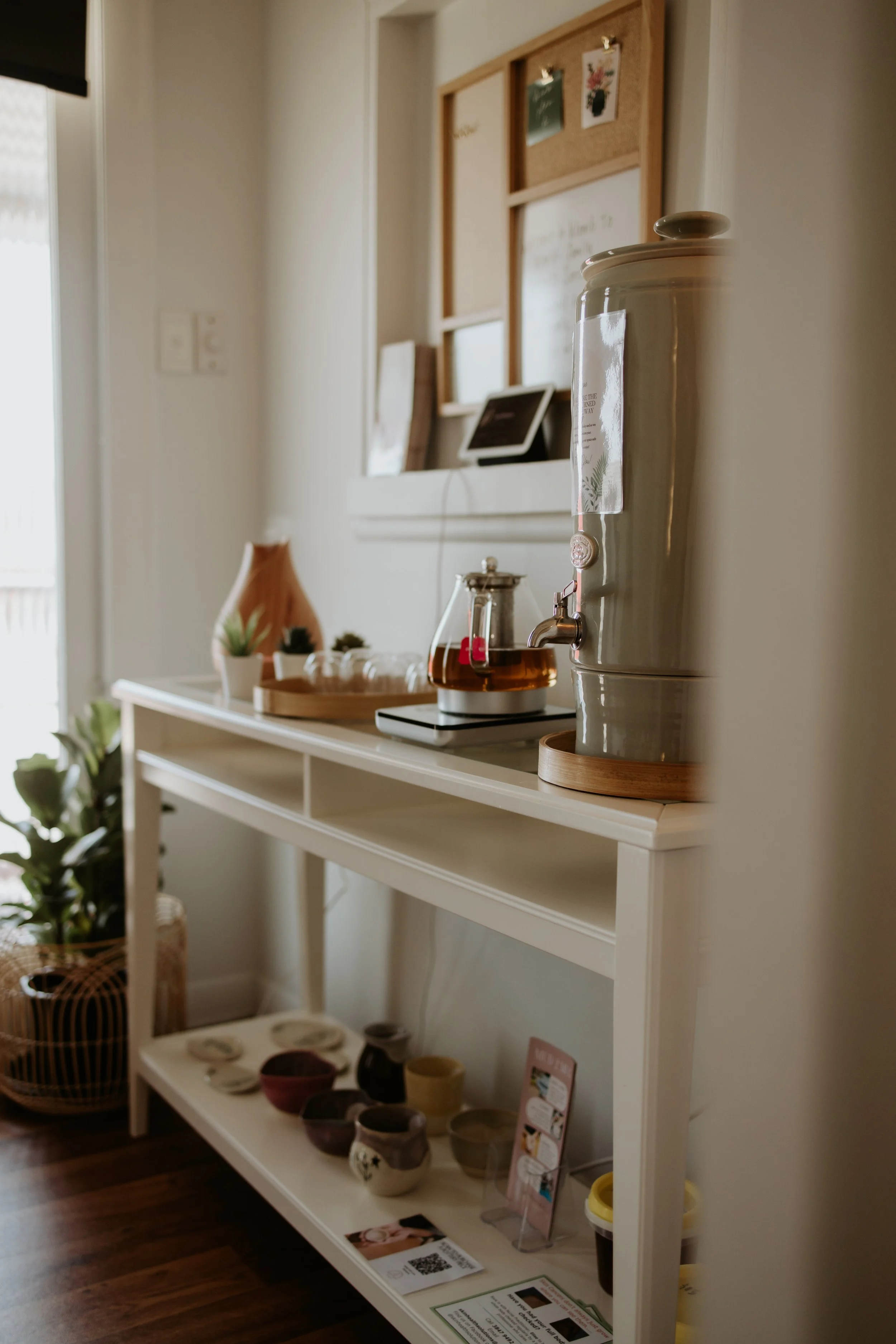 Home coffee bar with white table, large ceramic dispenser, teapot, potted plants, and various pottery mugs; bulletin board and electronic device on wall.