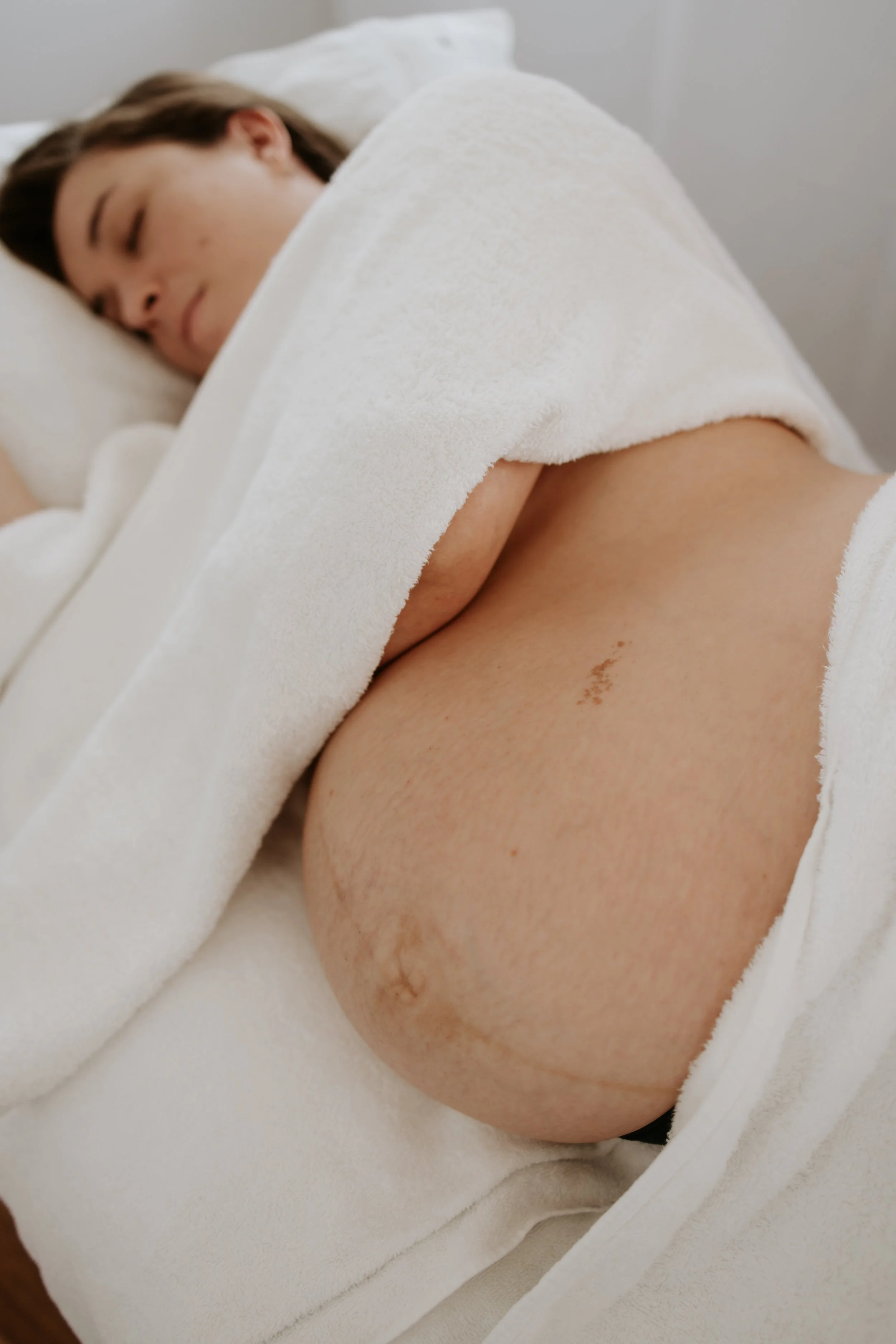 Pregnant woman lying on a bed, partly covered with a white towel, with a visible pregnant belly.
