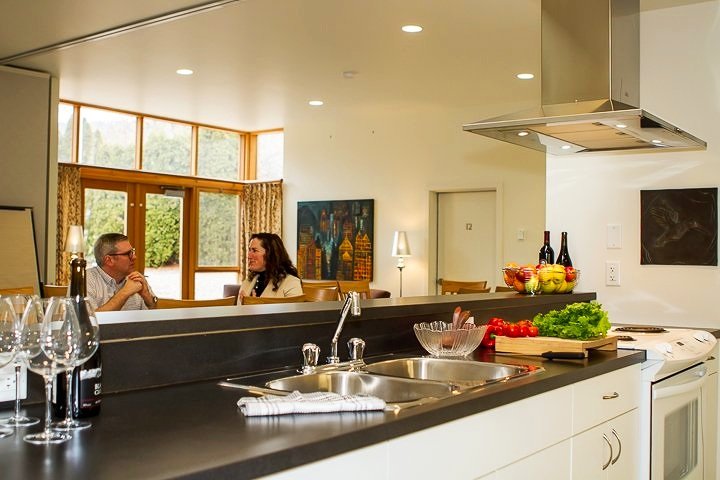 A modern kitchen with a black countertop, a double sink, and various items including wine glasses, a wine bottle, fresh fruits, vegetables, and bottles of wine. In the background, two people are sitting at a dining table near large windows.