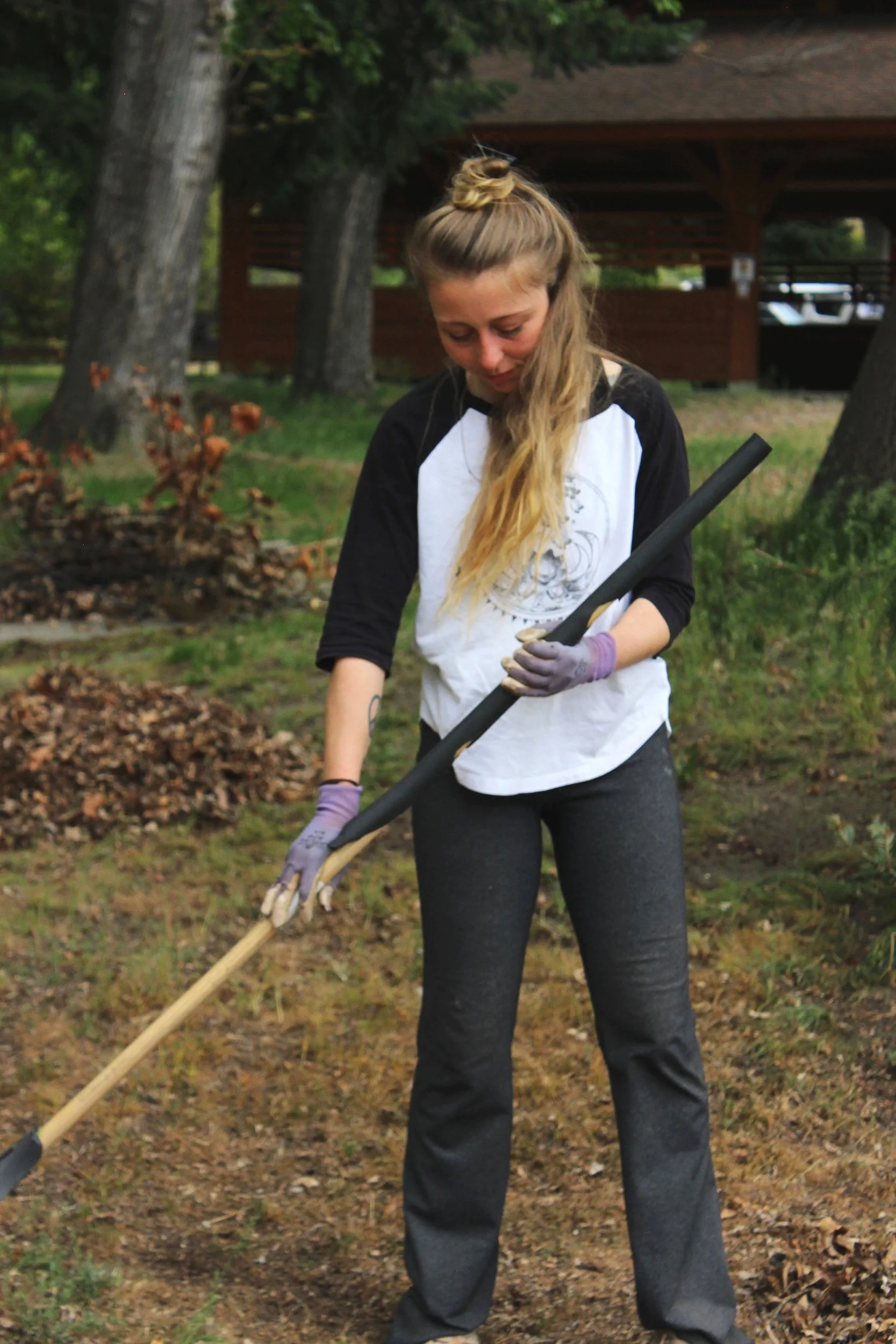 A woman with long blonde hair tied up in a bun, wearing a white and black shirt, gray pants, and purple gloves, is raking leaves in a yard surrounded by trees.