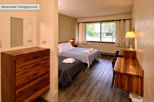 Hotel room at Naramata Campground with two beds, a large window, a wooden dresser, and a wooden desk with a lamp.