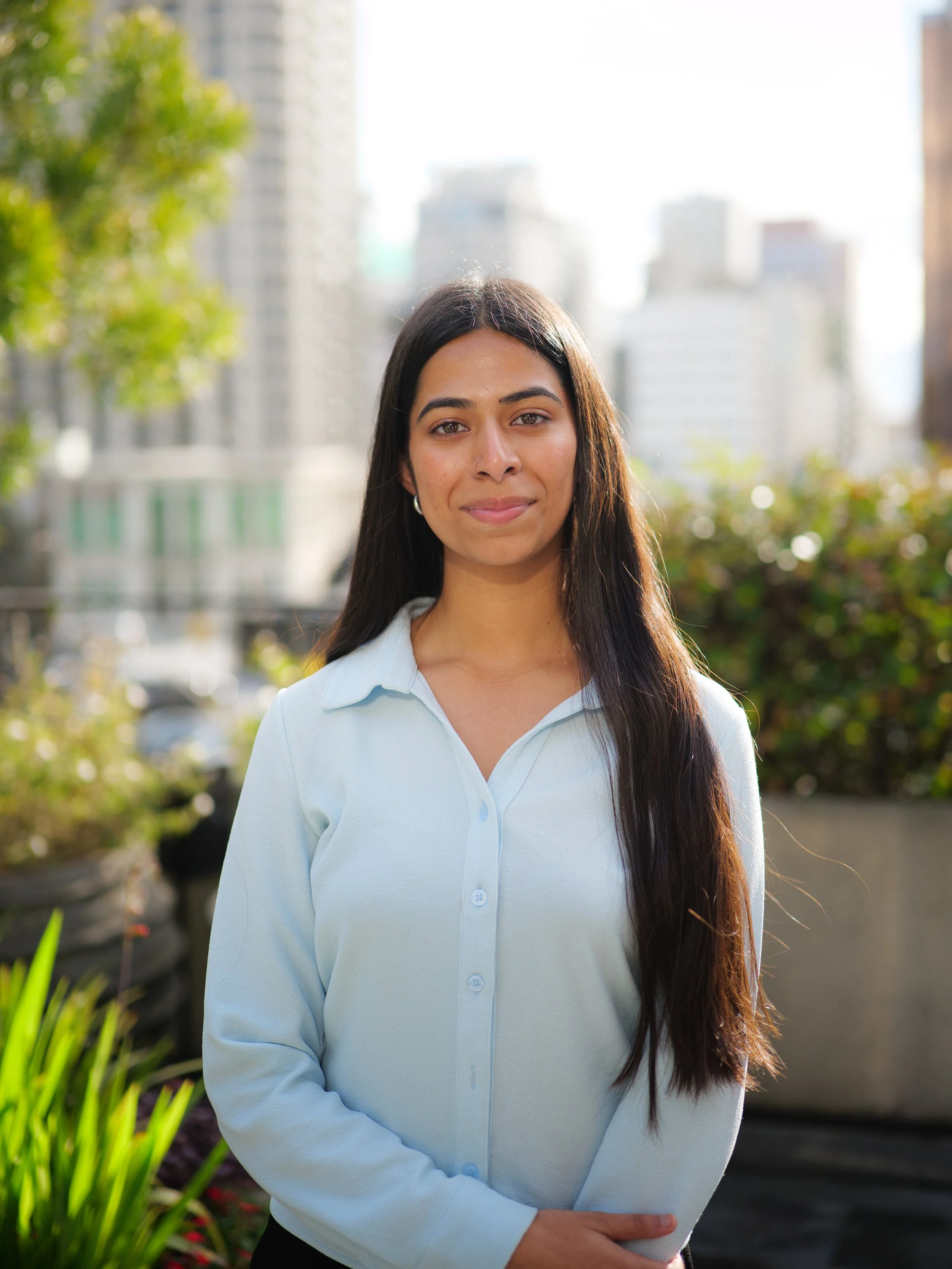 A woman with long dark hair, wearing a black and gray T-shirt, standing outdoors with arms crossed, smiling softly, against a beige stone wall background.