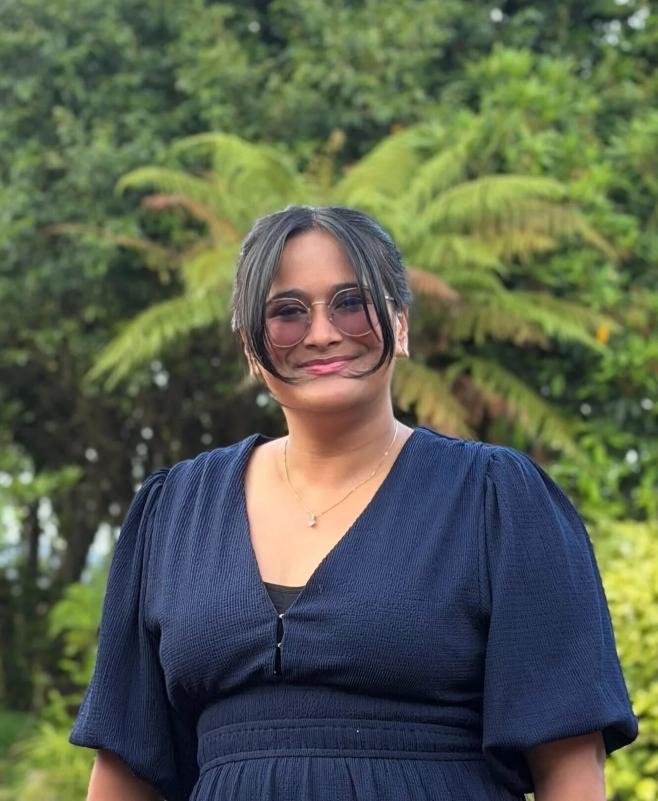 A woman with short dark hair, wearing glasses, a navy blue dress, and a gold necklace, smiling outdoors with green trees in the background.
