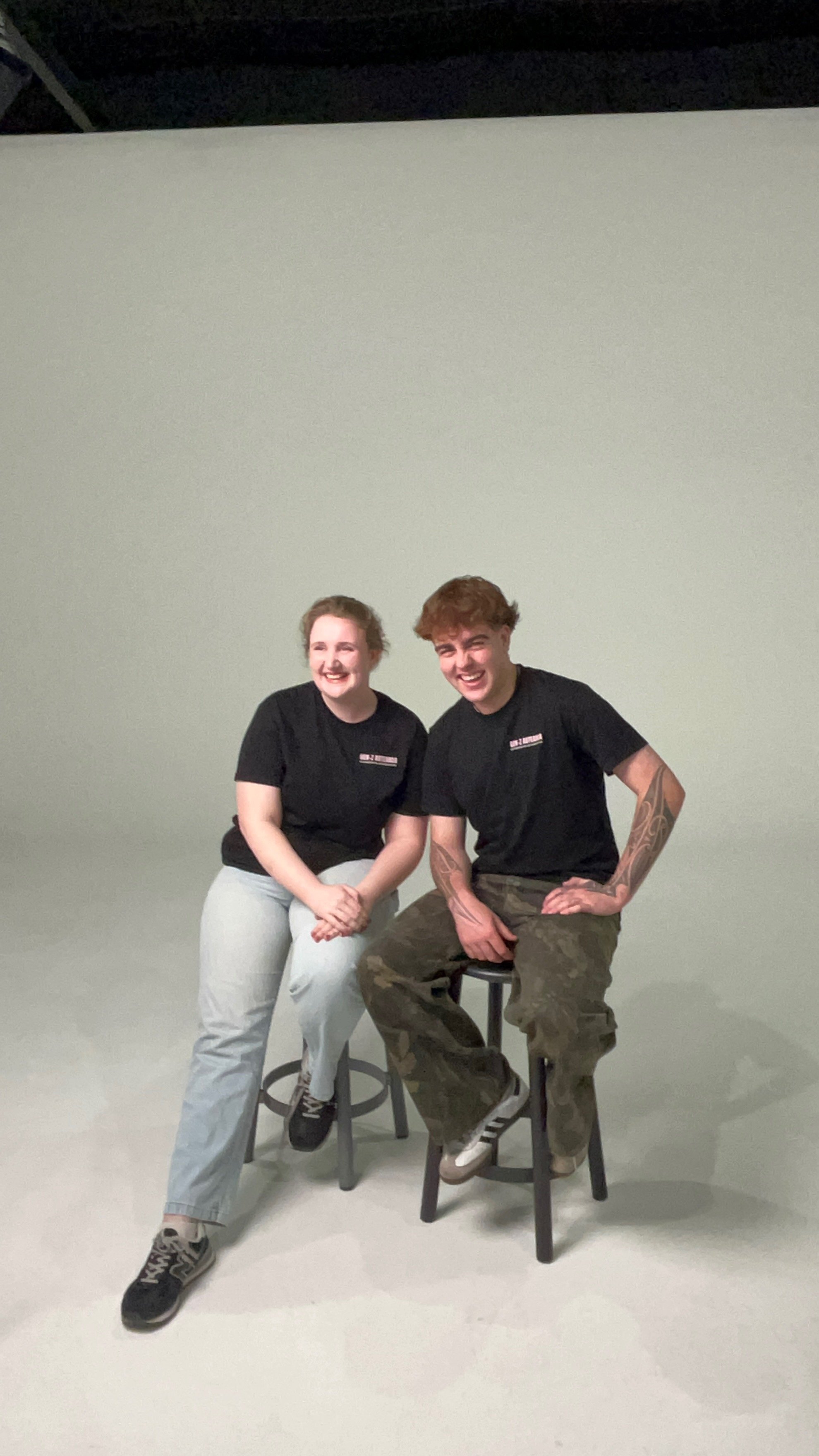 Two smiling young people, a woman and a man, sitting on a stool in front of a plain backdrop, wearing black t-shirts and casual pants.