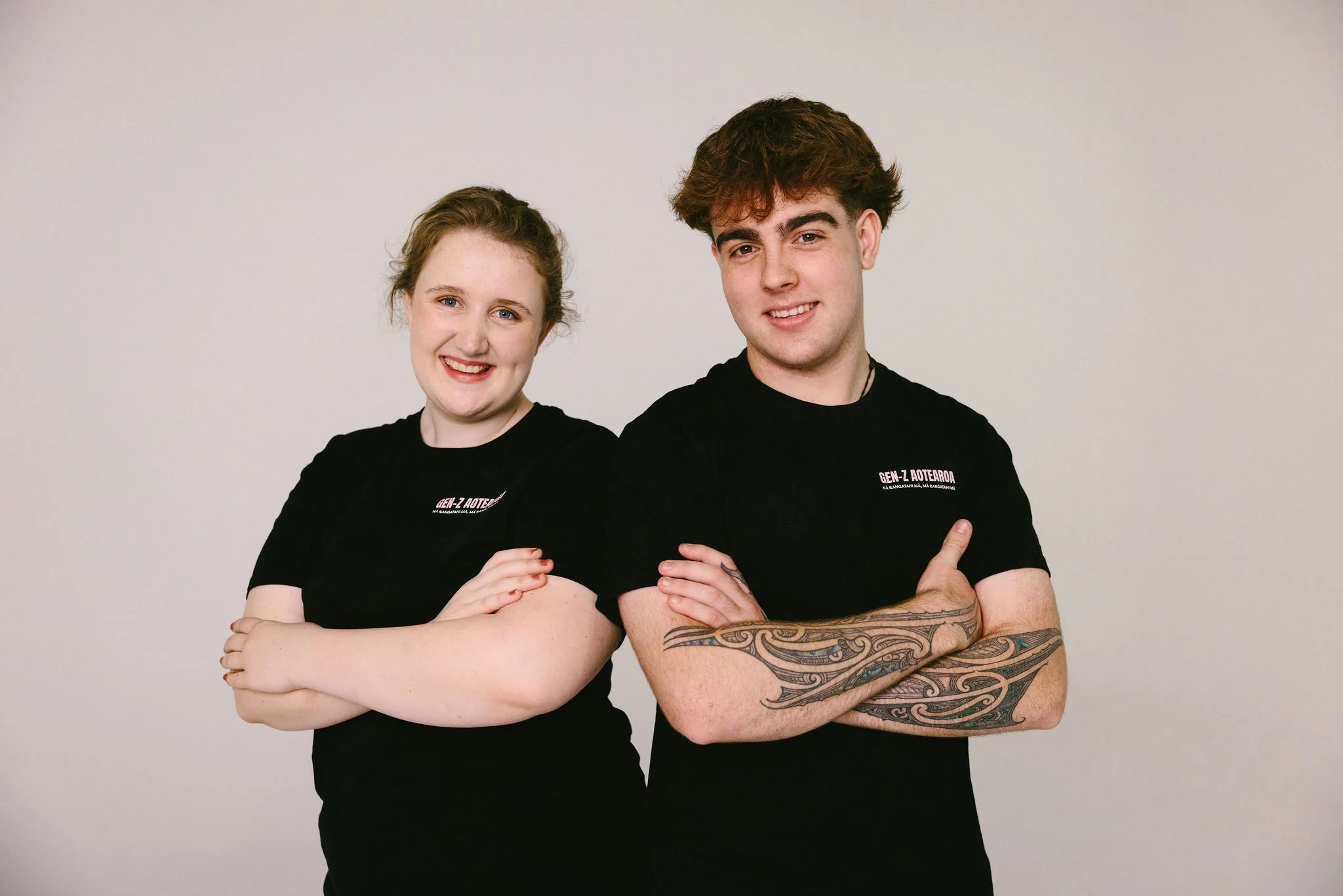 Two young adults, a woman and a man, standing side by side with their arms crossed, smiling in front of a plain white background, wearing black t-shirts.