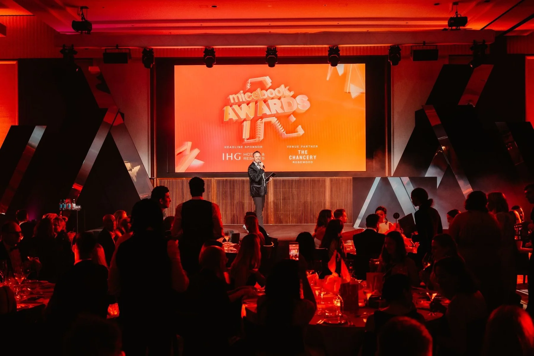 Host on stage at the Micebook Awards 2026 in front of a large branded screen and a full room of dinner guests