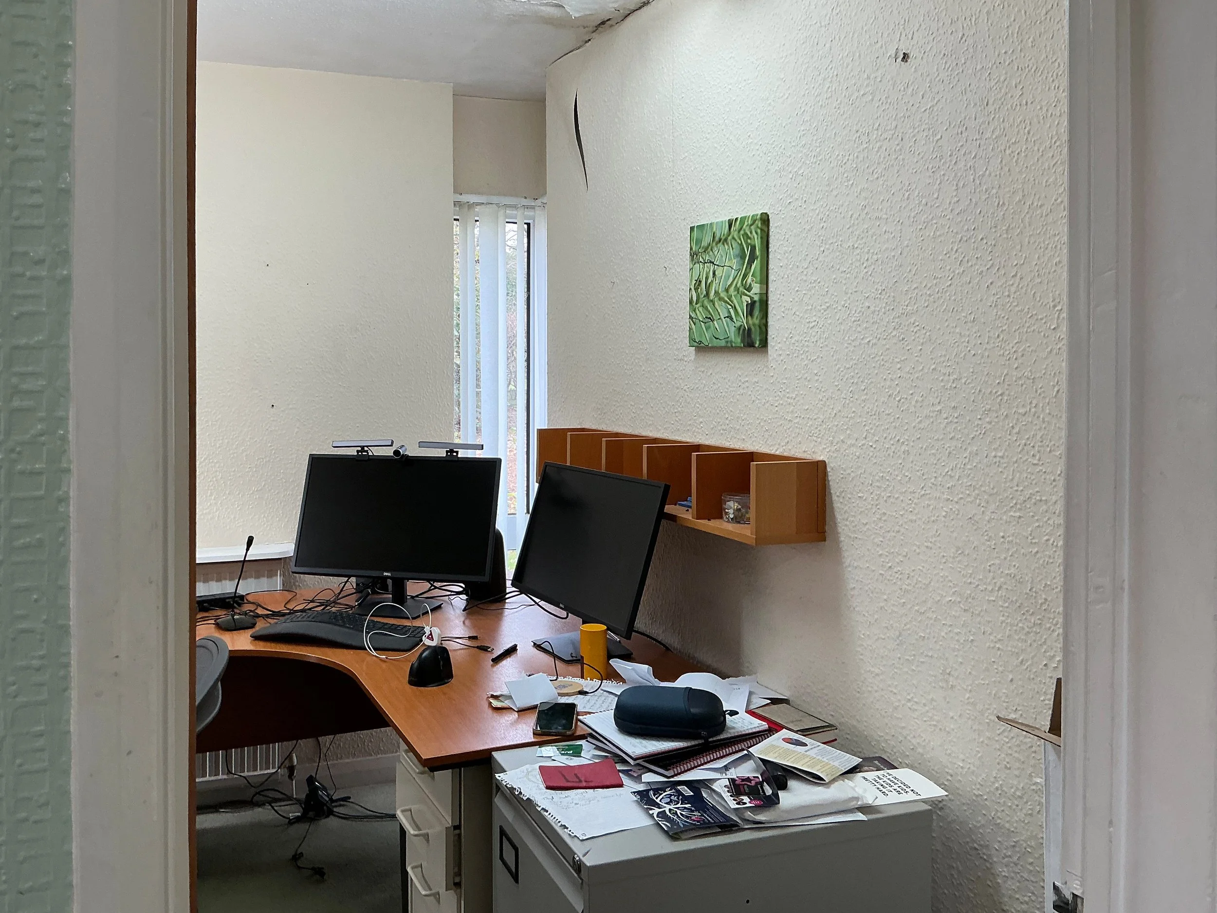 A photo through a doorway of a home office. It has a large corner desk, on it sit two monitors and lots of messy papers